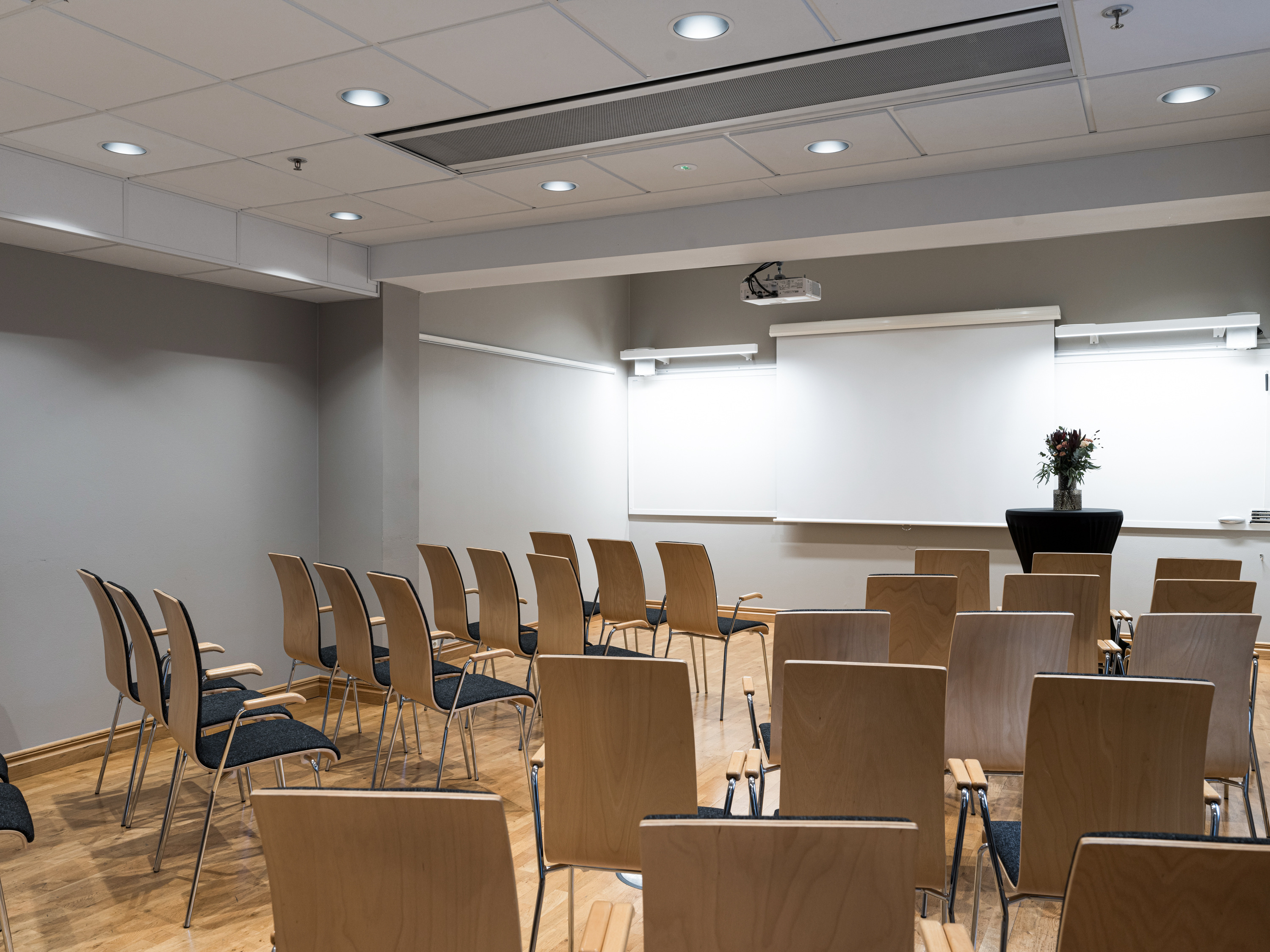 Modern conference room with wooden chairs, a projector, a large screen, and a floral arrangement, ready for a presentation or meeting.