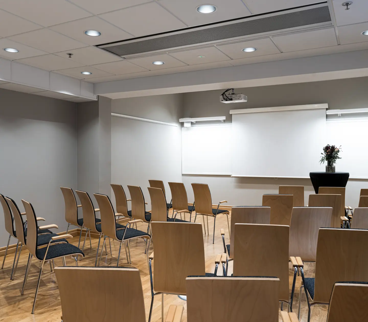 Modern conference room with wooden chairs, a projector, a large screen, and a floral arrangement, ready for a presentation or meeting.