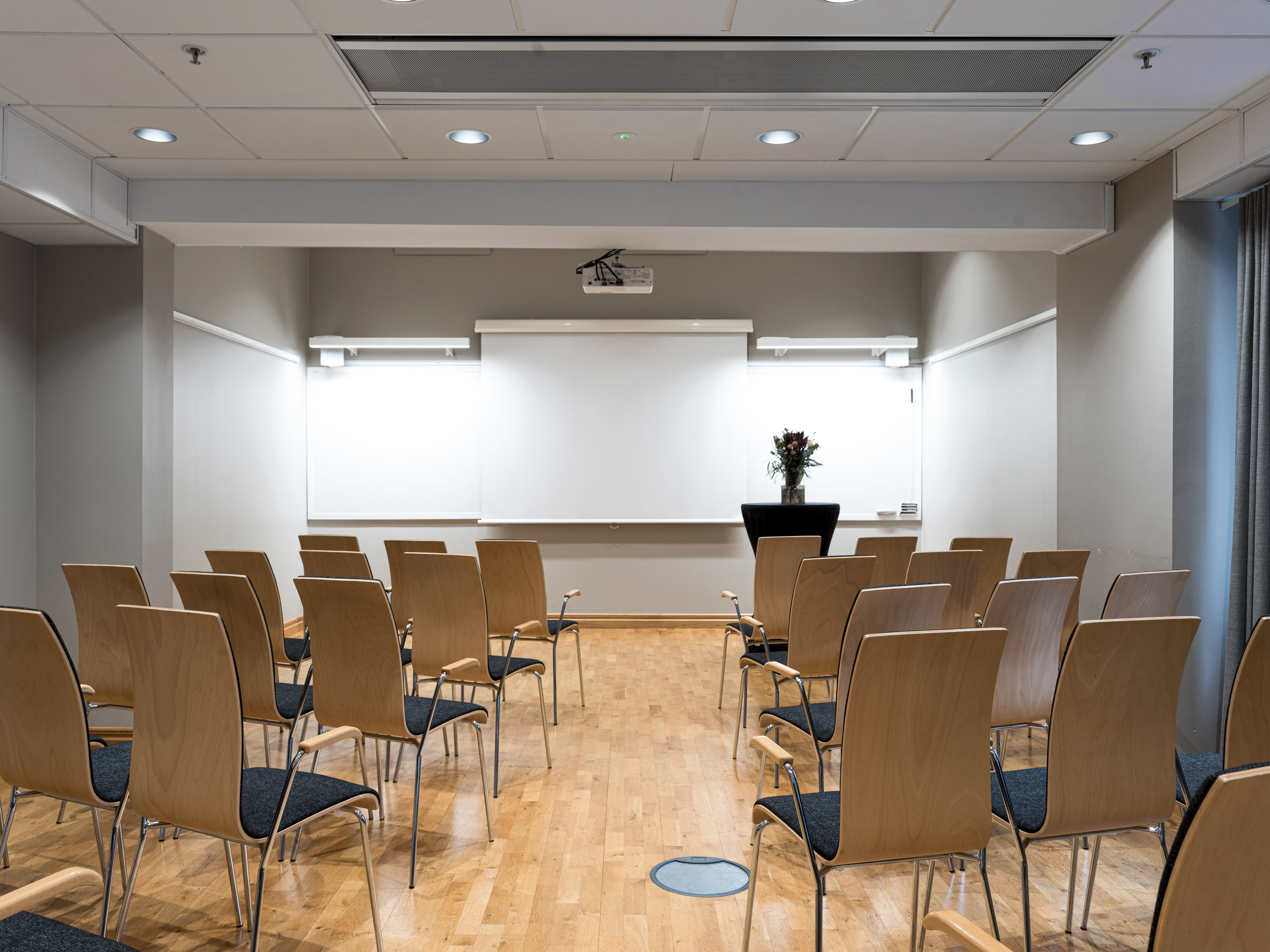 Modern conference room with rows of empty wooden chairs facing a projector screen and whiteboards, ready for a presentation or meeting.