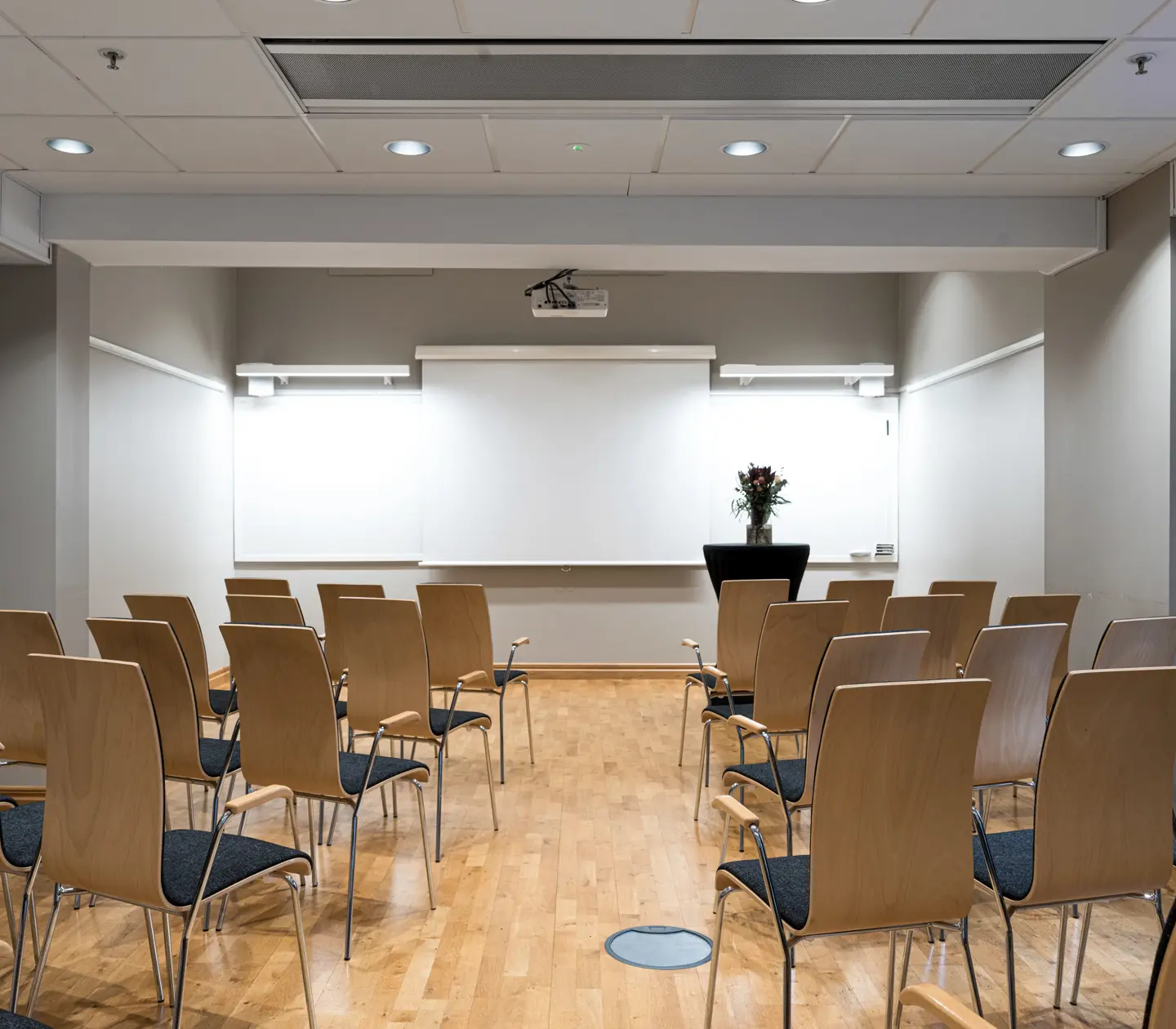 Modern conference room with rows of empty wooden chairs facing a projector screen and whiteboards, ready for a presentation or meeting.