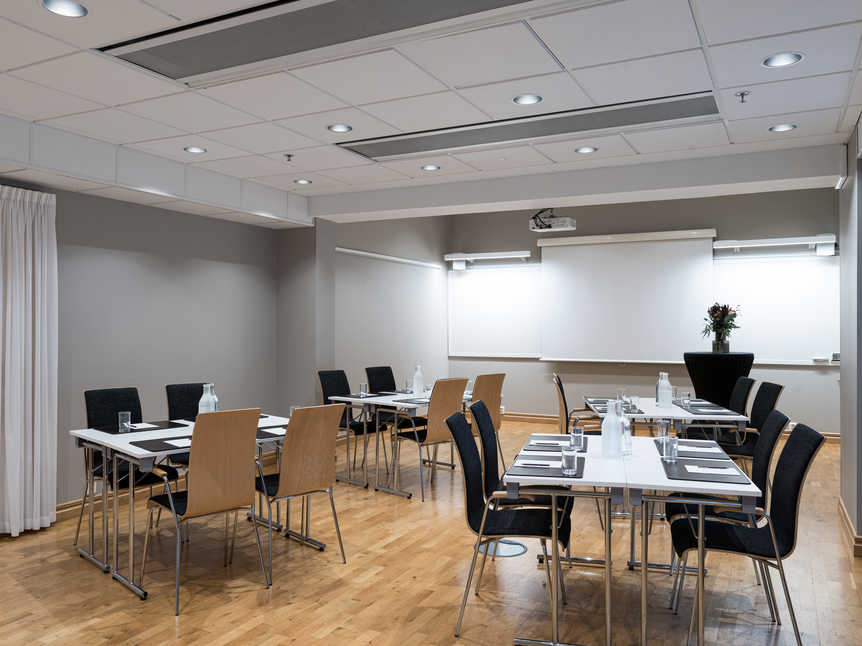 Modern conference room with beige walls and wood flooring, featuring set tables with black and beige chairs, notepads, and water bottles, ready for a business meeting or presentation.