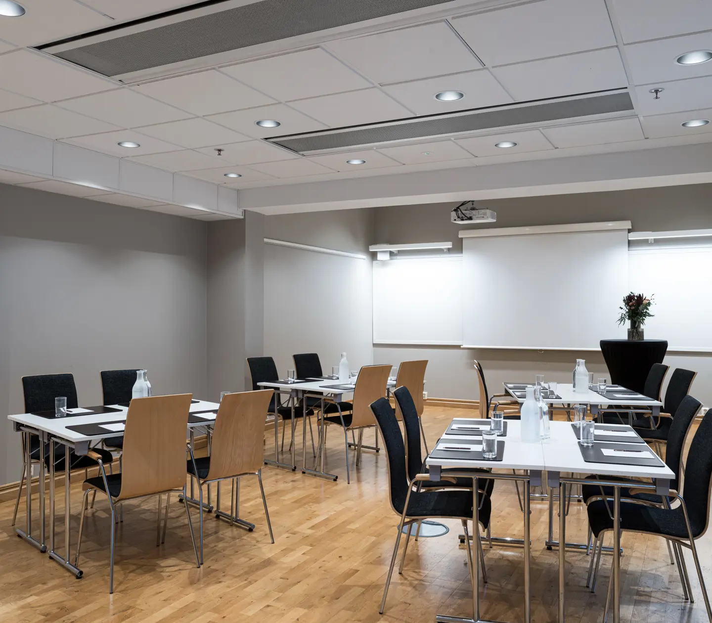 Modern conference room with beige walls and wood flooring, featuring set tables with black and beige chairs, notepads, and water bottles, ready for a business meeting or presentation.