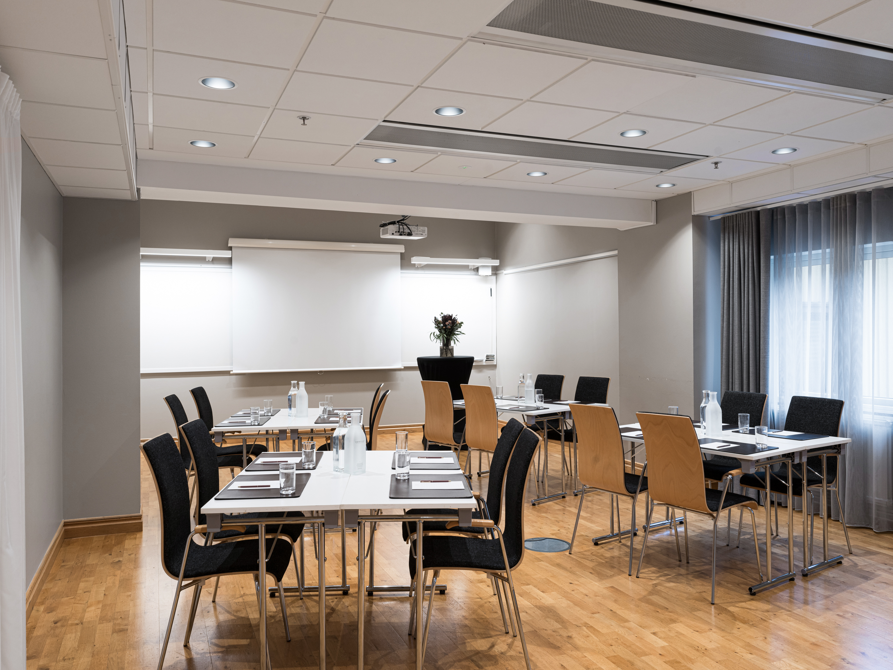 Modern conference rooms with projector screens, wooden tables, and black chairs, prepared for meetings with water bottles and glasses.