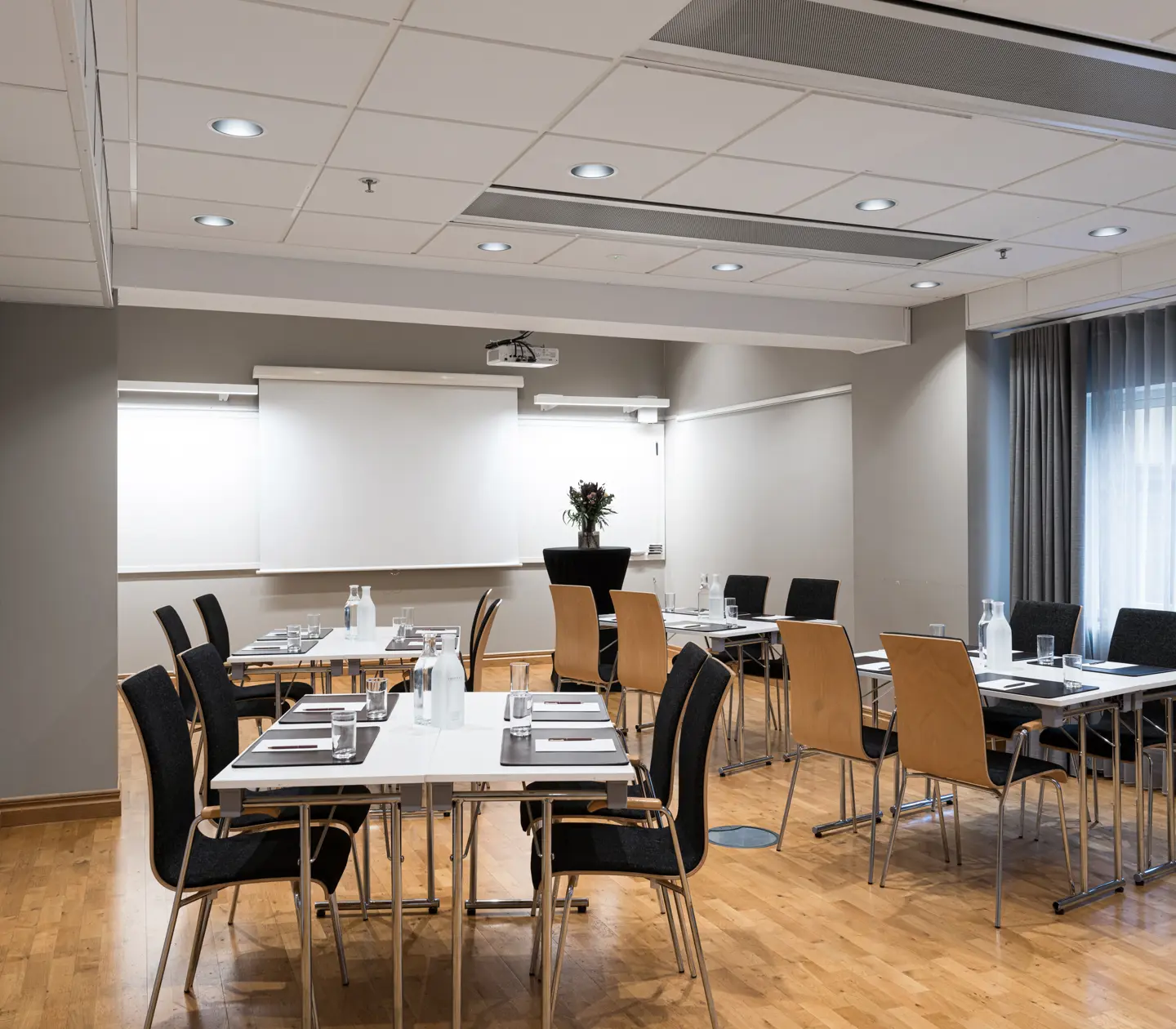Modern conference rooms with projector screens, wooden tables, and black chairs, prepared for meetings with water bottles and glasses.