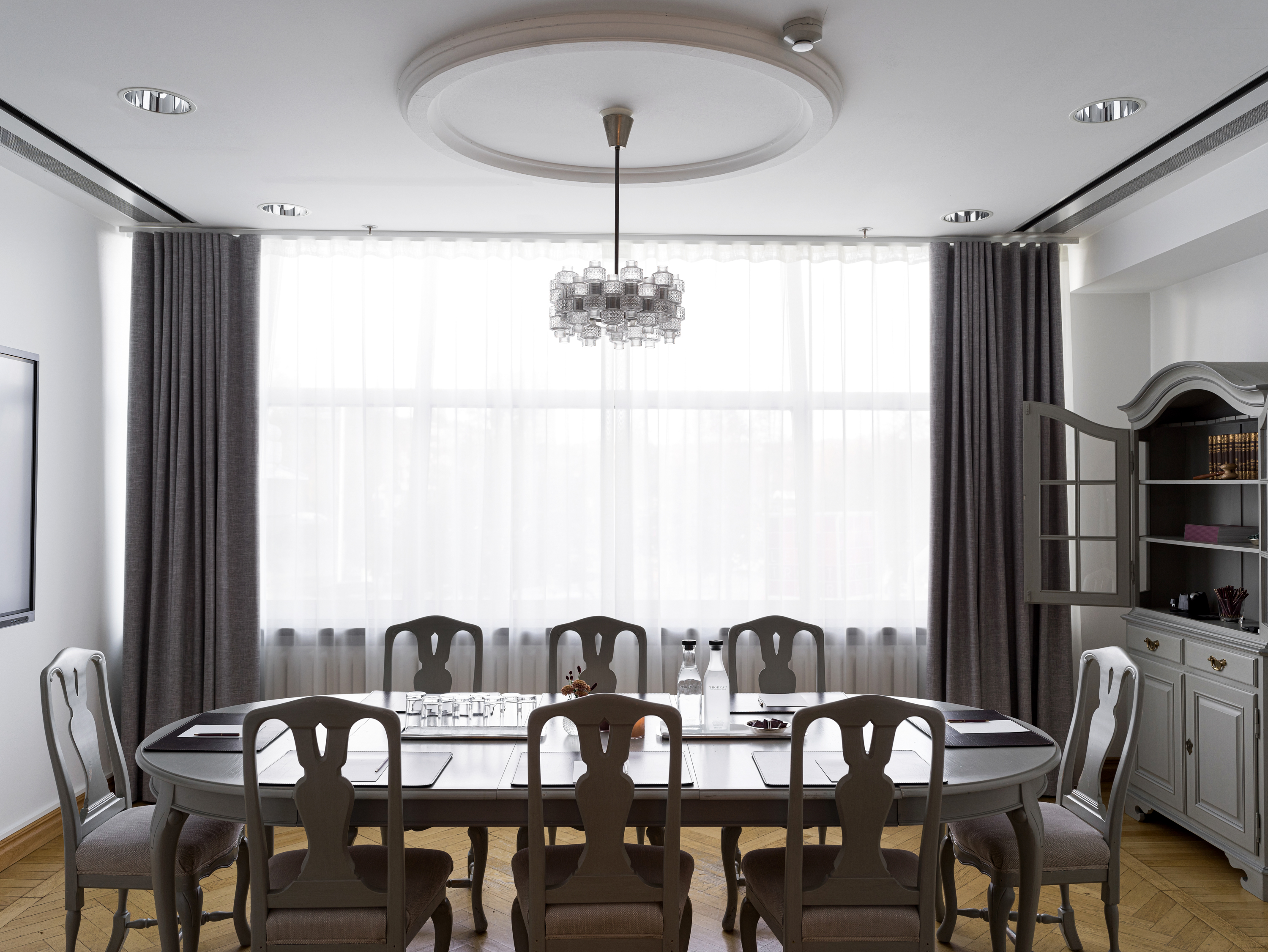 Elegant dining room with a large oval table, vintage chairs, a modern chandelier, and natural light flowing through floor-to-ceiling curtains.