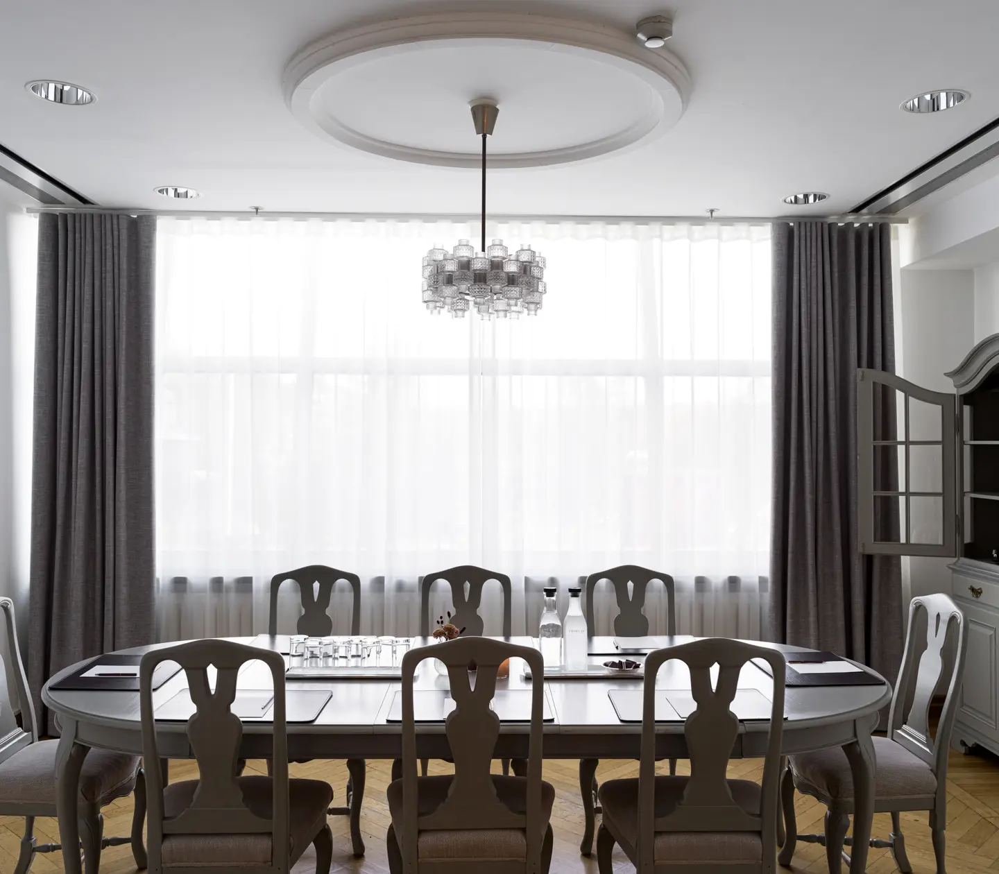 Elegant dining room with a large oval table, vintage chairs, a modern chandelier, and natural light flowing through floor-to-ceiling curtains.