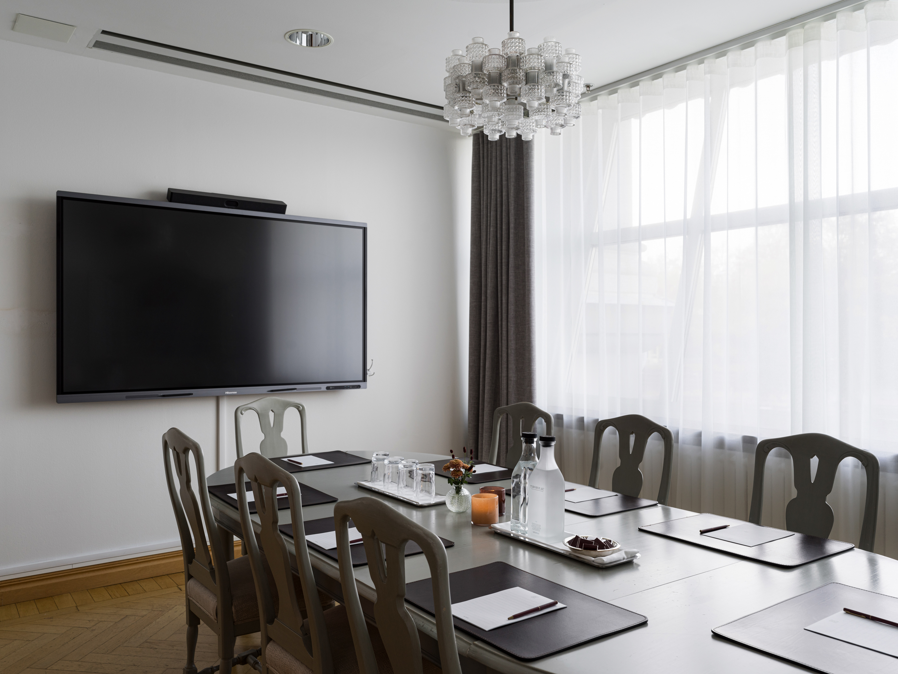 Modern conference room with a large wall-mounted TV, elegant chandelier, and a table set for meetings. Features include chairs, notebooks, and water bottles, positioned near a large window with sheer curtains.