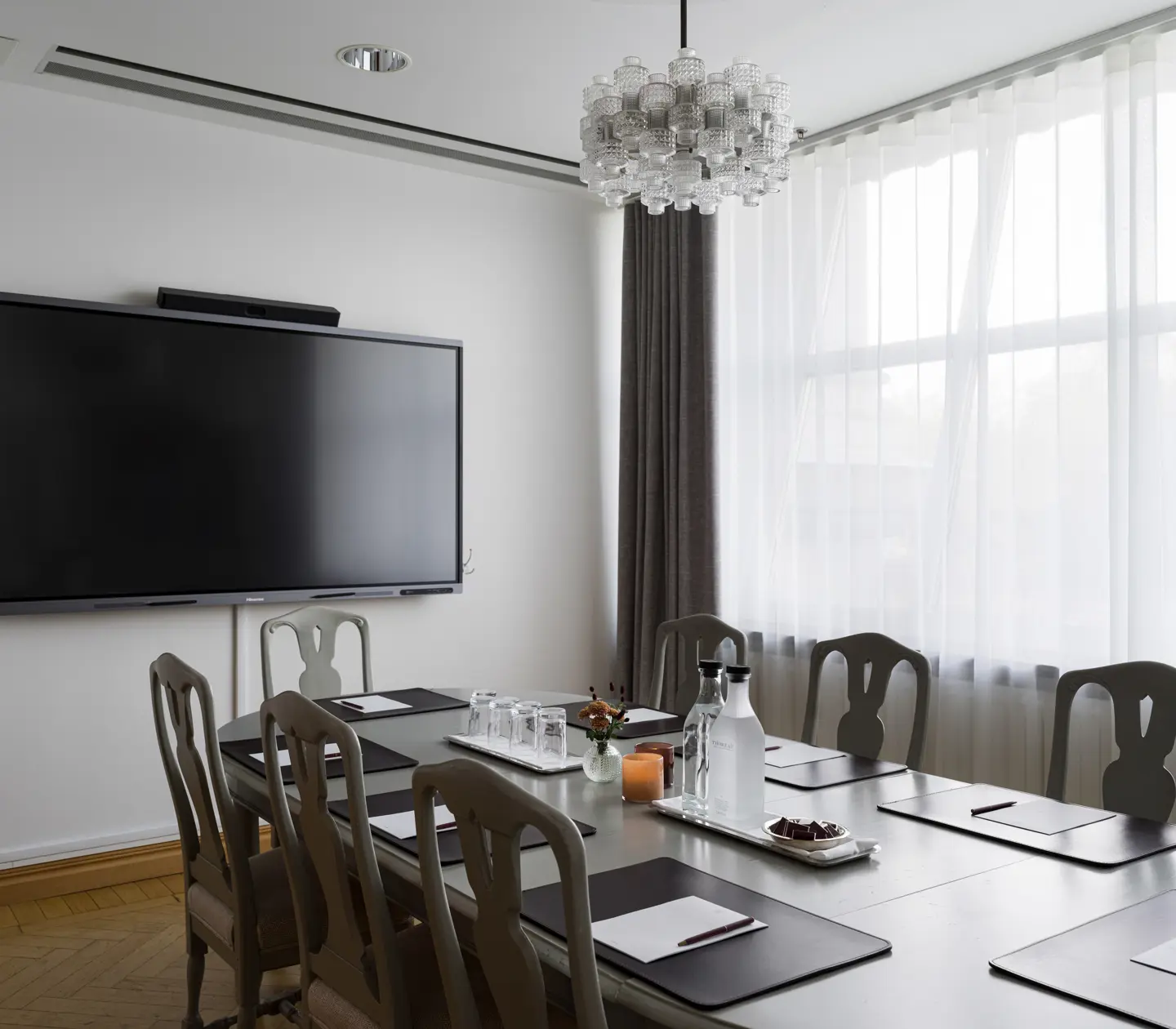 Modern conference room with a large wall-mounted TV, elegant chandelier, and a table set for meetings. Features include chairs, notebooks, and water bottles, positioned near a large window with sheer curtains.