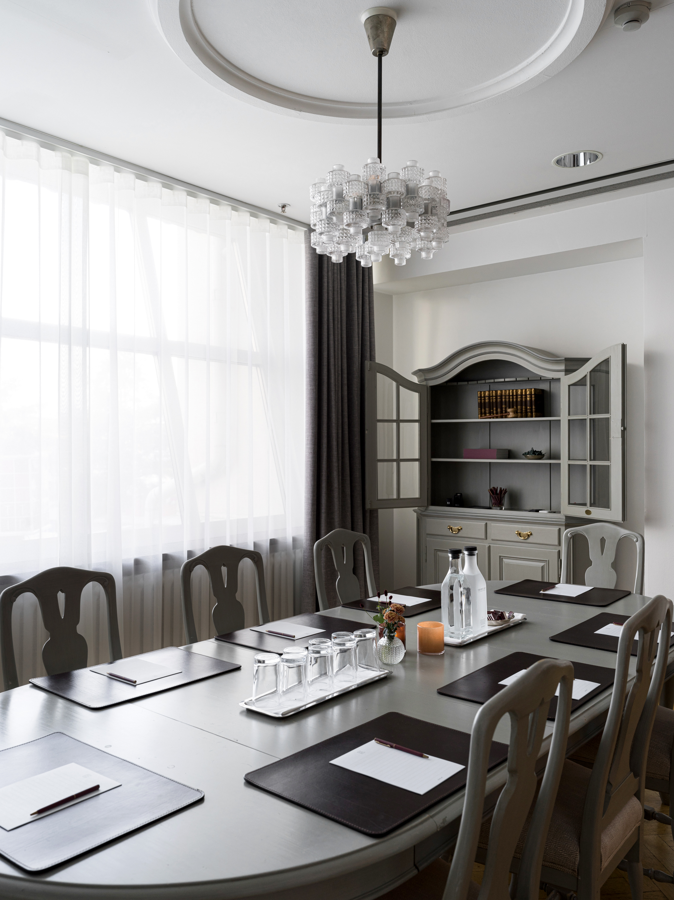 Conference room with a large oval table, chairs, and place settings including notepads and water bottles, situated near a window with sheer curtains and featuring a decorative cabinet in the background.