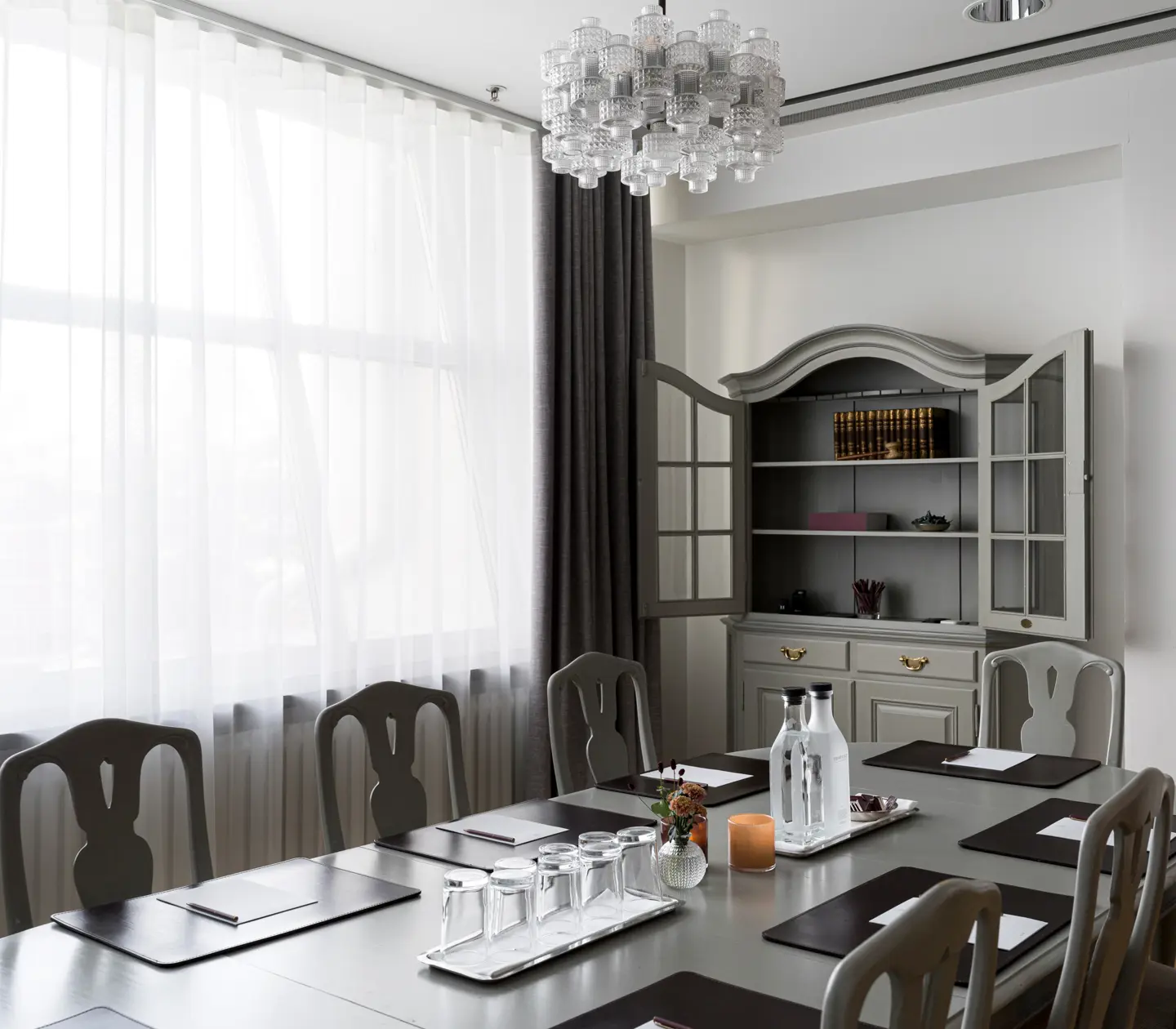 Conference room with a large oval table, chairs, and place settings including notepads and water bottles, situated near a window with sheer curtains and featuring a decorative cabinet in the background.