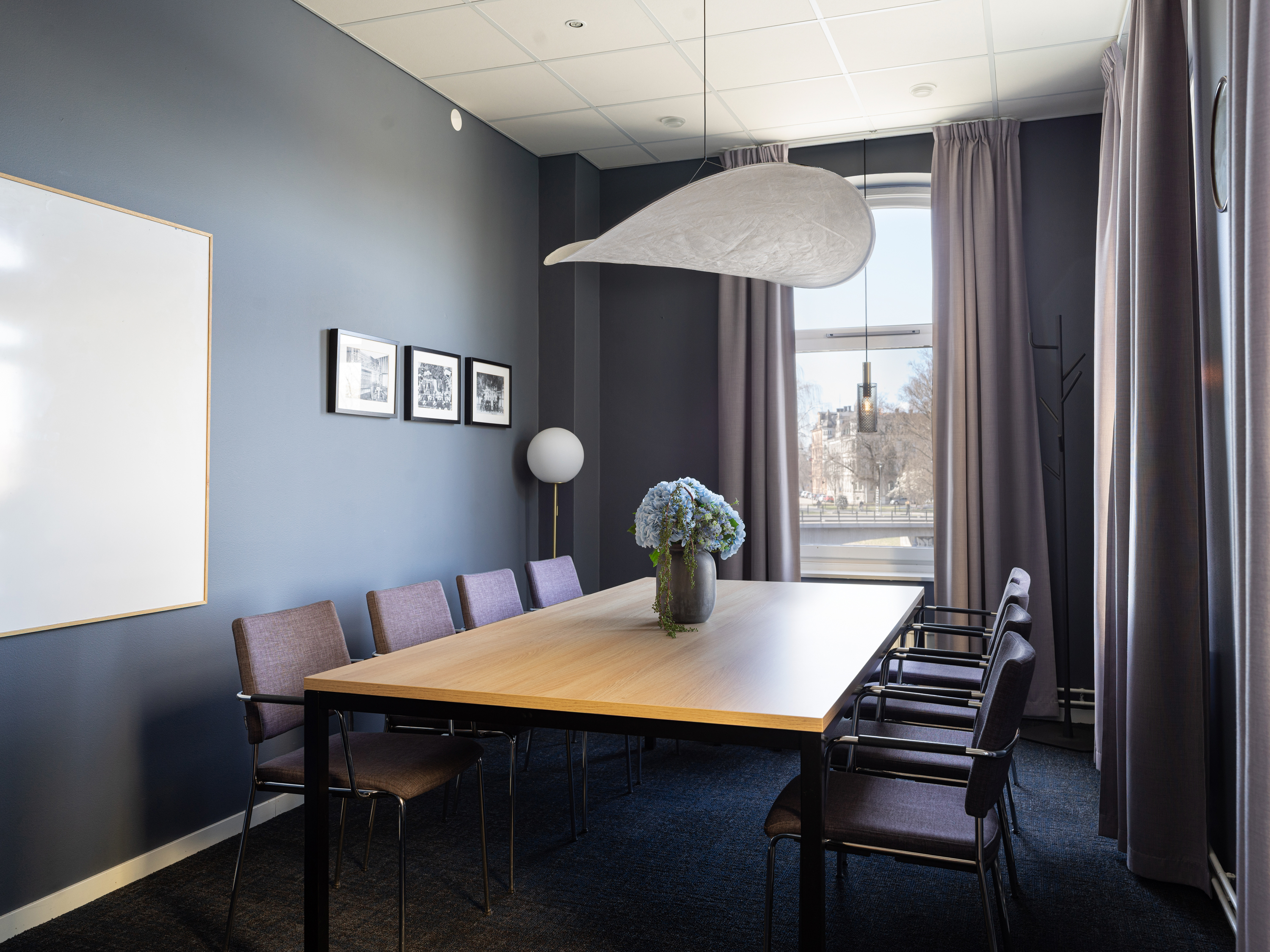 Modern conference room with a wooden table, surrounded by chairs and decorated with a vase of blue flowers. The room features a blue accent wall, framed artwork, and large window with gray curtains, creating a professional atmosphere.