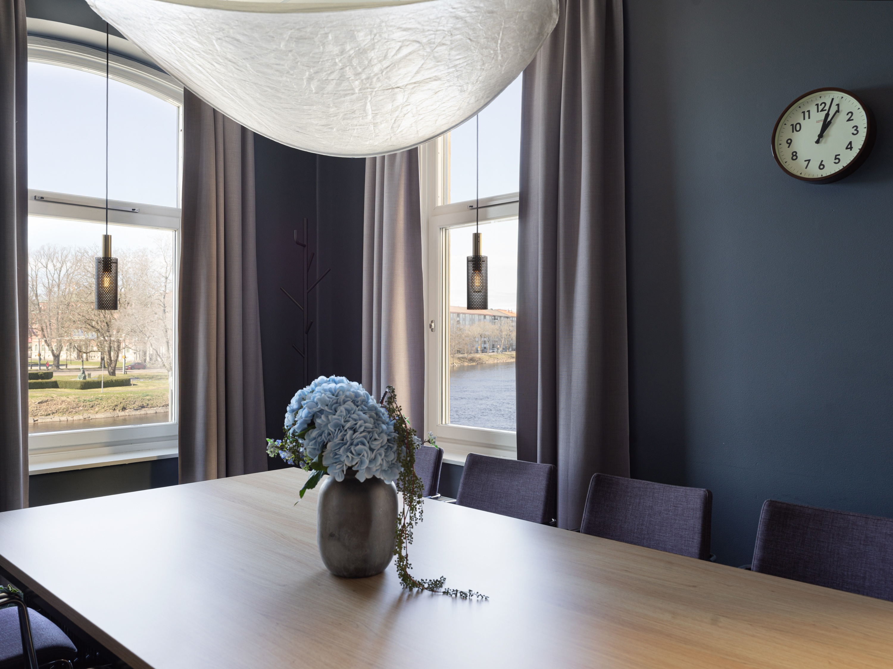 Modern dining room with a wooden table, vase of blue flowers, and dark curtains. Large windows reveal a river view. Pendant lights and a clock enhance the decor.