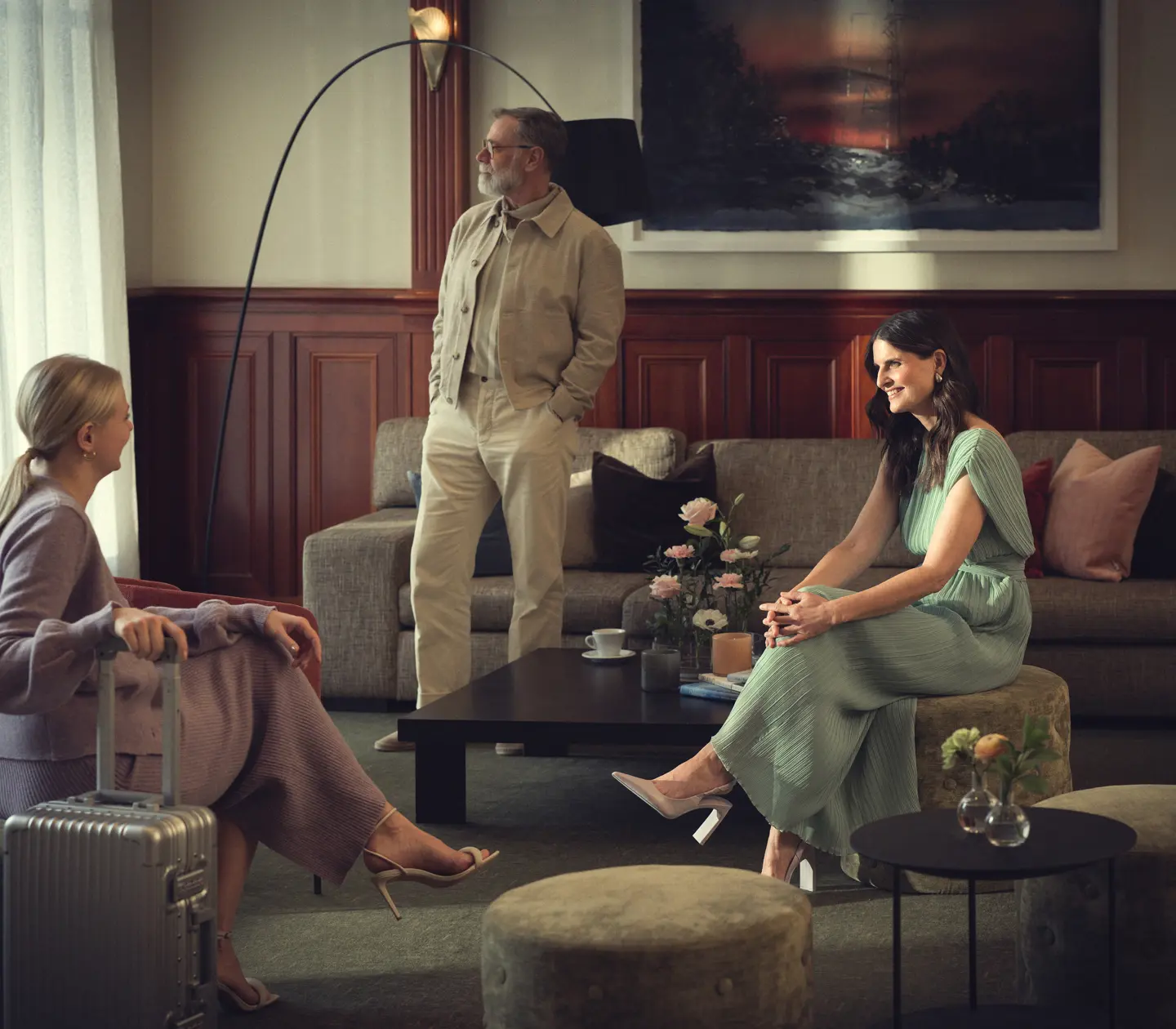 Elegant lobby setting with three people in contemporary clothing, furnished with sofa, armchairs and a still life on the table.