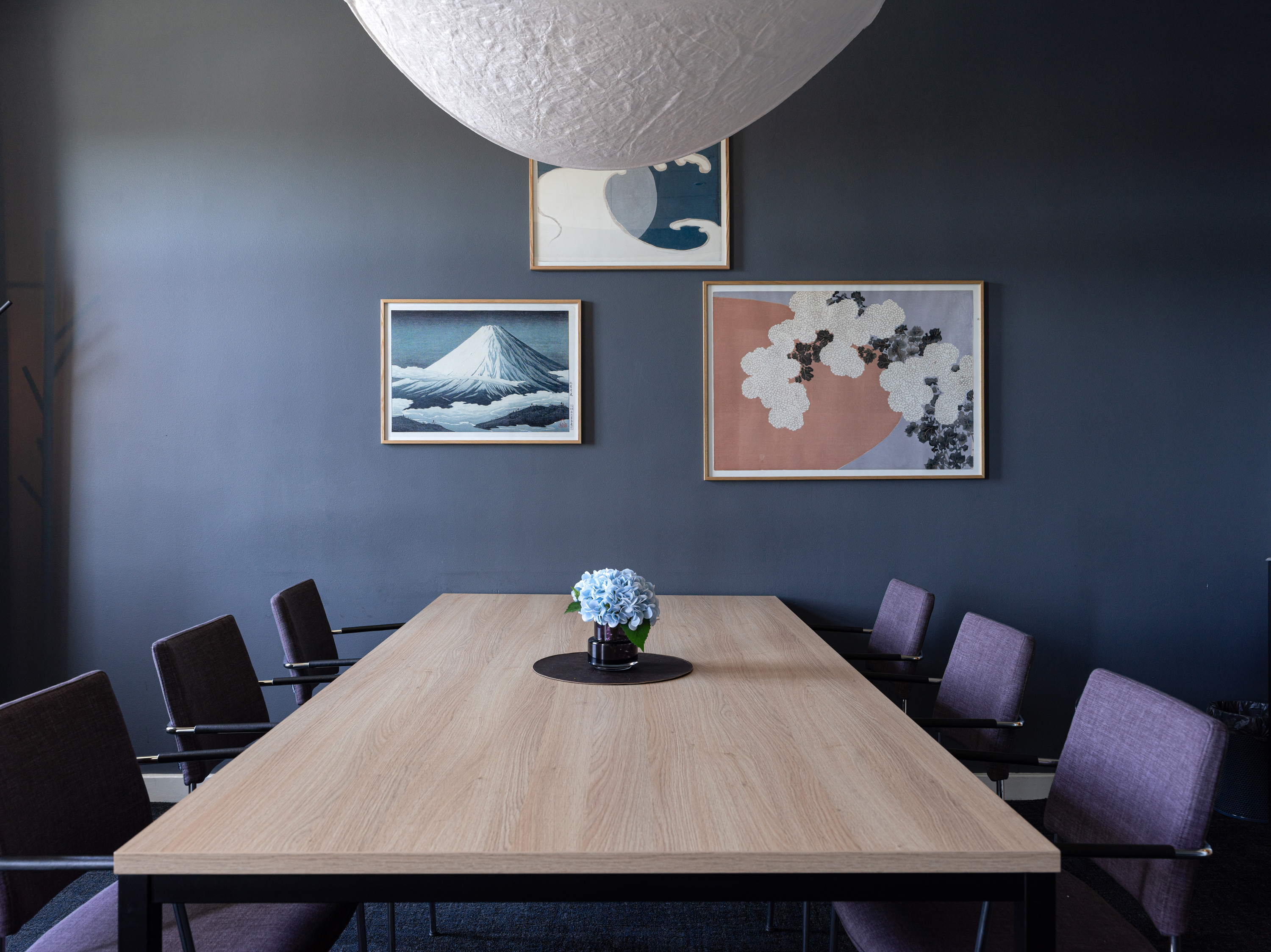 Minimalist conference room with a large wooden table, eight purple chairs, and a central flower arrangement under a modern white pendant light. Three art pieces with nature-themed designs adorn the dark walls.