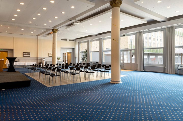 Large room with blue carpet, lined up chairs and large windows