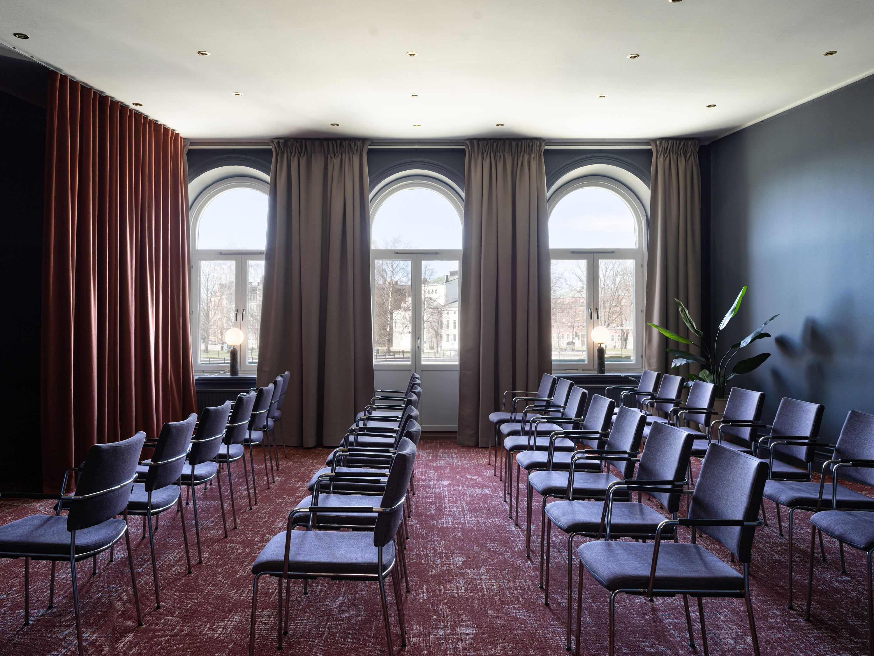 Conference room with arched windows, burgundy curtains, and rows of empty chairs on a red carpet, ready for an event.