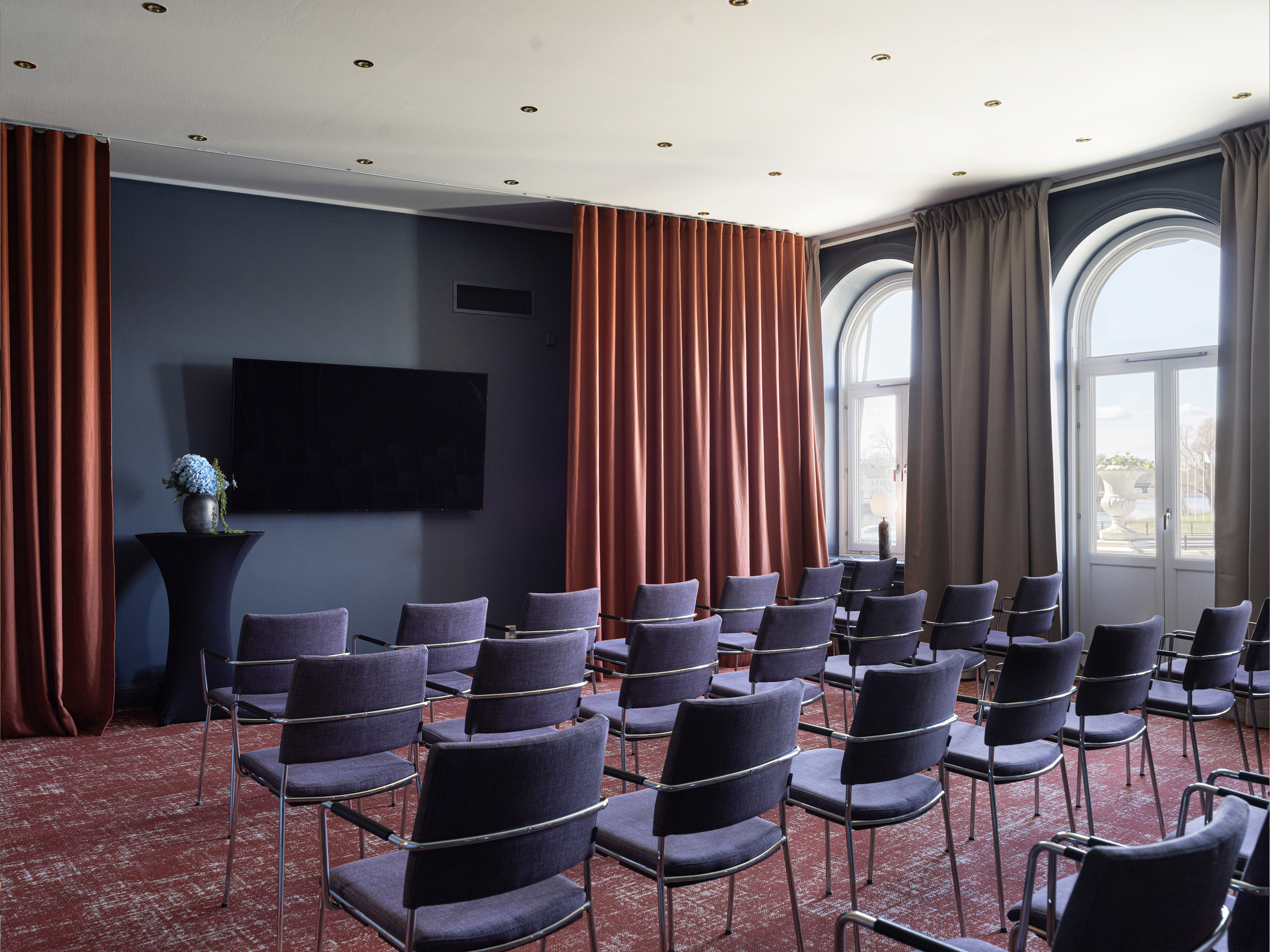 Conference room setup with rows of empty chairs facing a large flat-screen television, framed by dark walls and large windows with red and gray curtains.