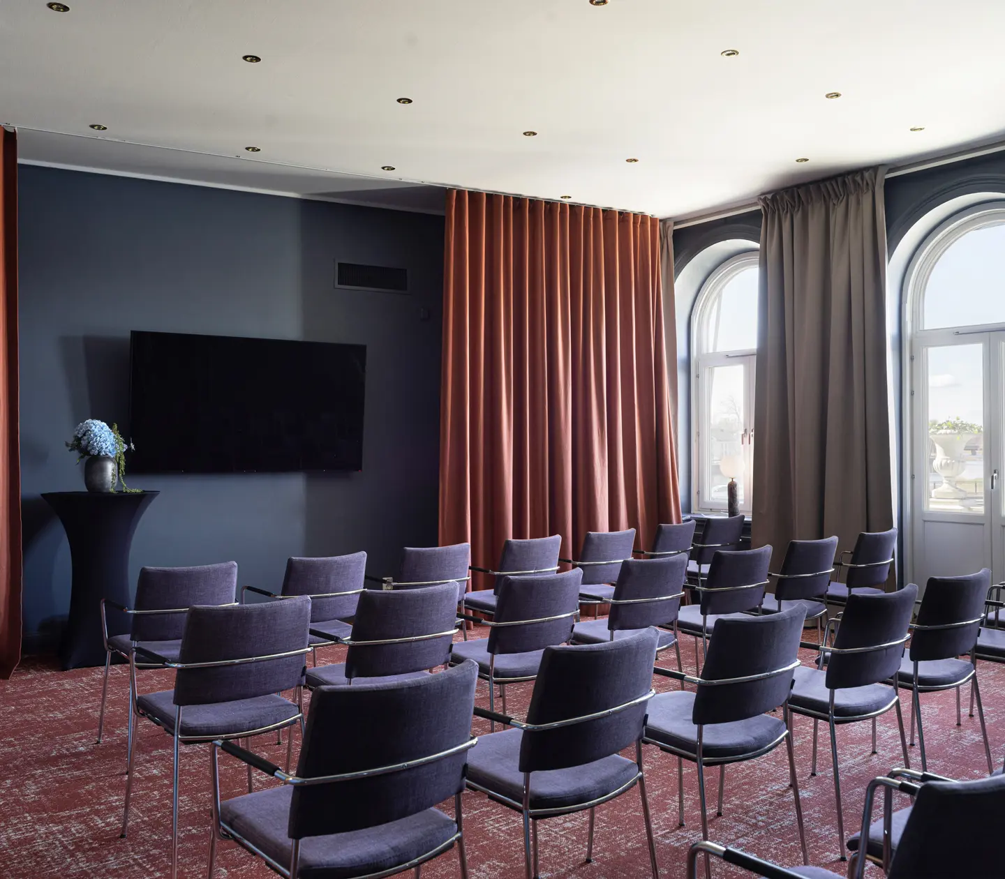 Konferensrum med stolar, mörkblå väggar, och stora fönster med gardiner.