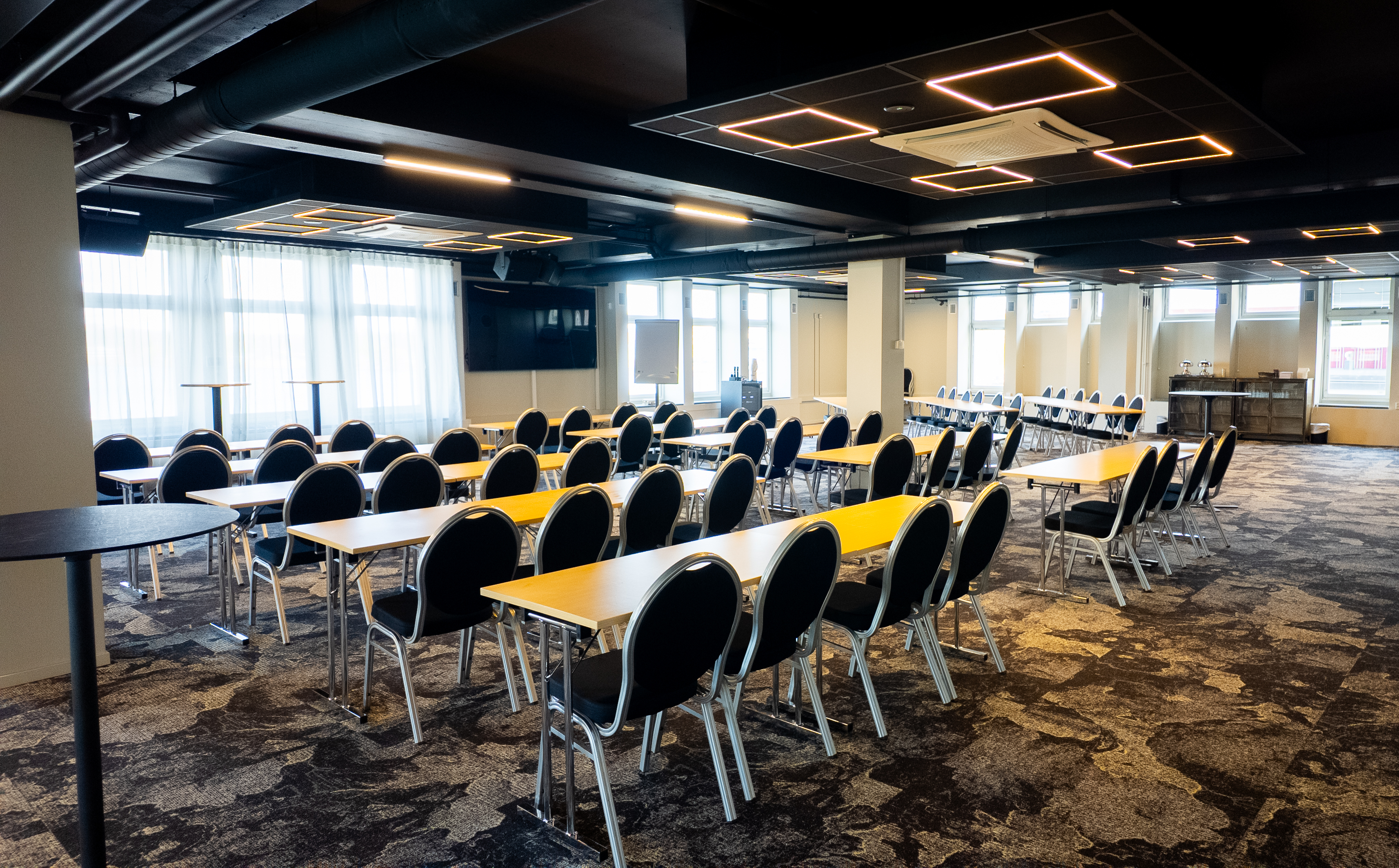 Spacious conference room with rows of tables and chairs, modern lighting, and large windows allowing natural light. 