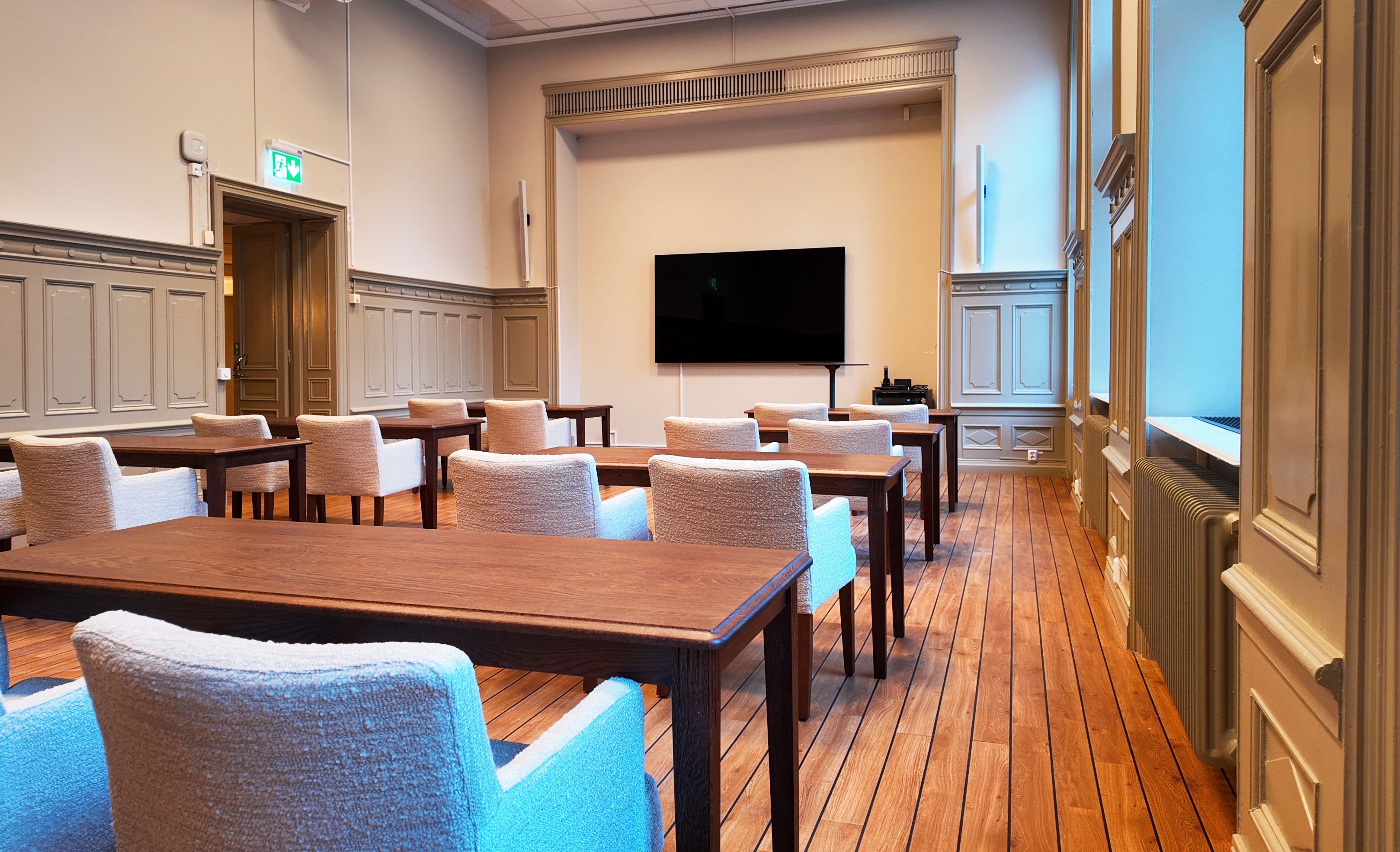 Modern conference room with wooden floors, upholstered chairs, and a large wall-mounted flat screen TV.