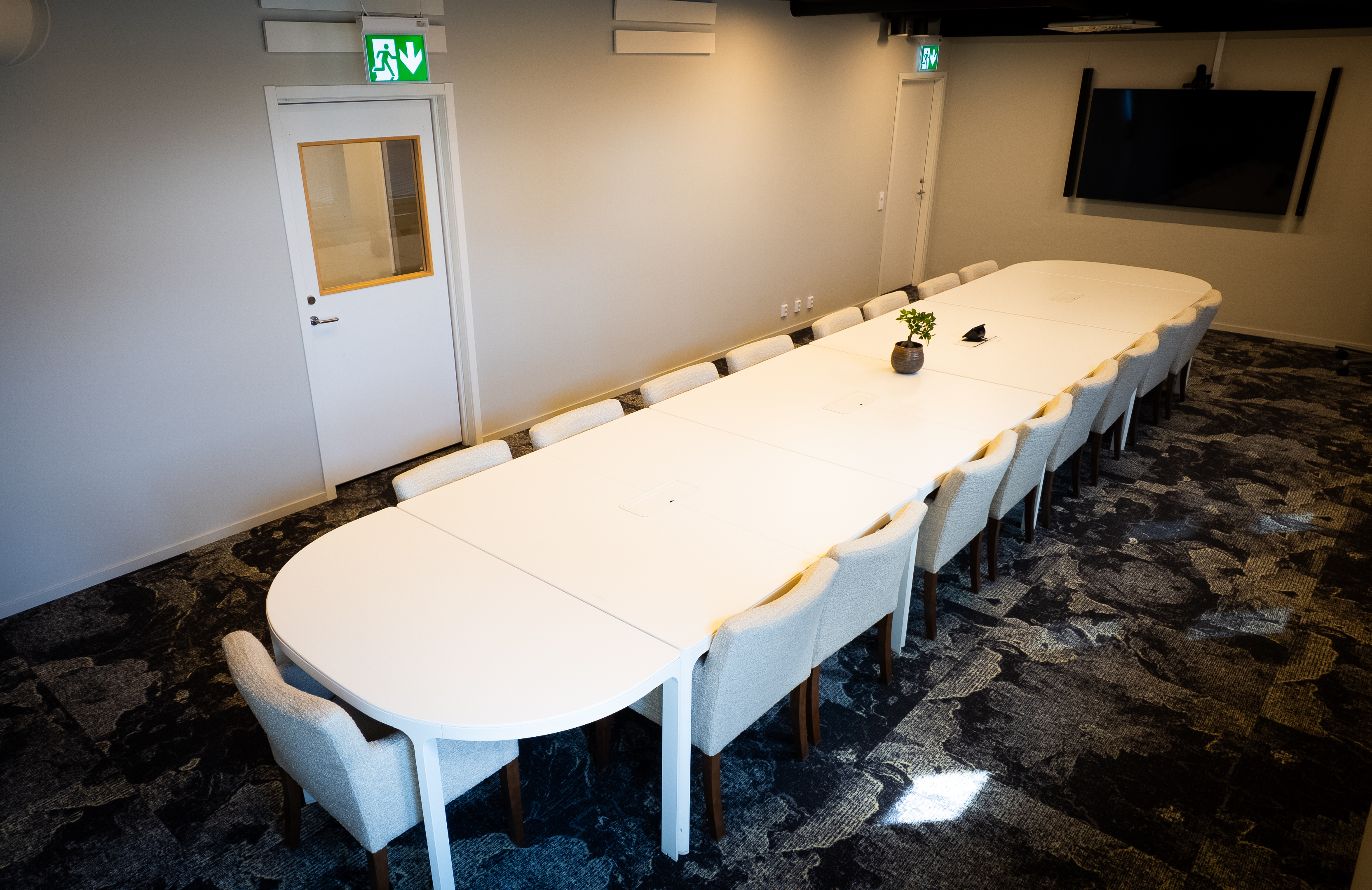 Modern conference room with a long white table surrounded by white chairs, featuring a wall-mounted screen and contemporary carpet flooring.