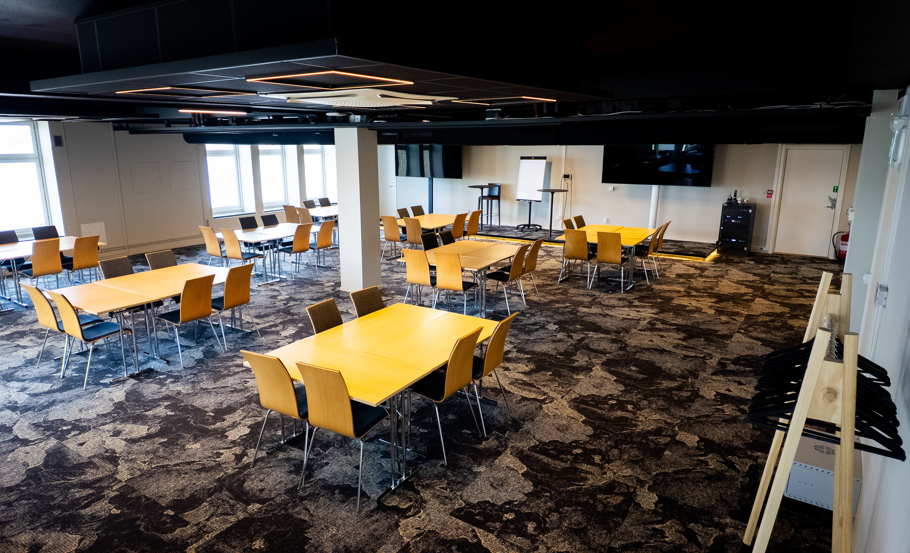 Spacious conference room with multiple tables and chairs, modern decor, large windows providing natural light, and presentation equipment including a flipchart and TV screens.