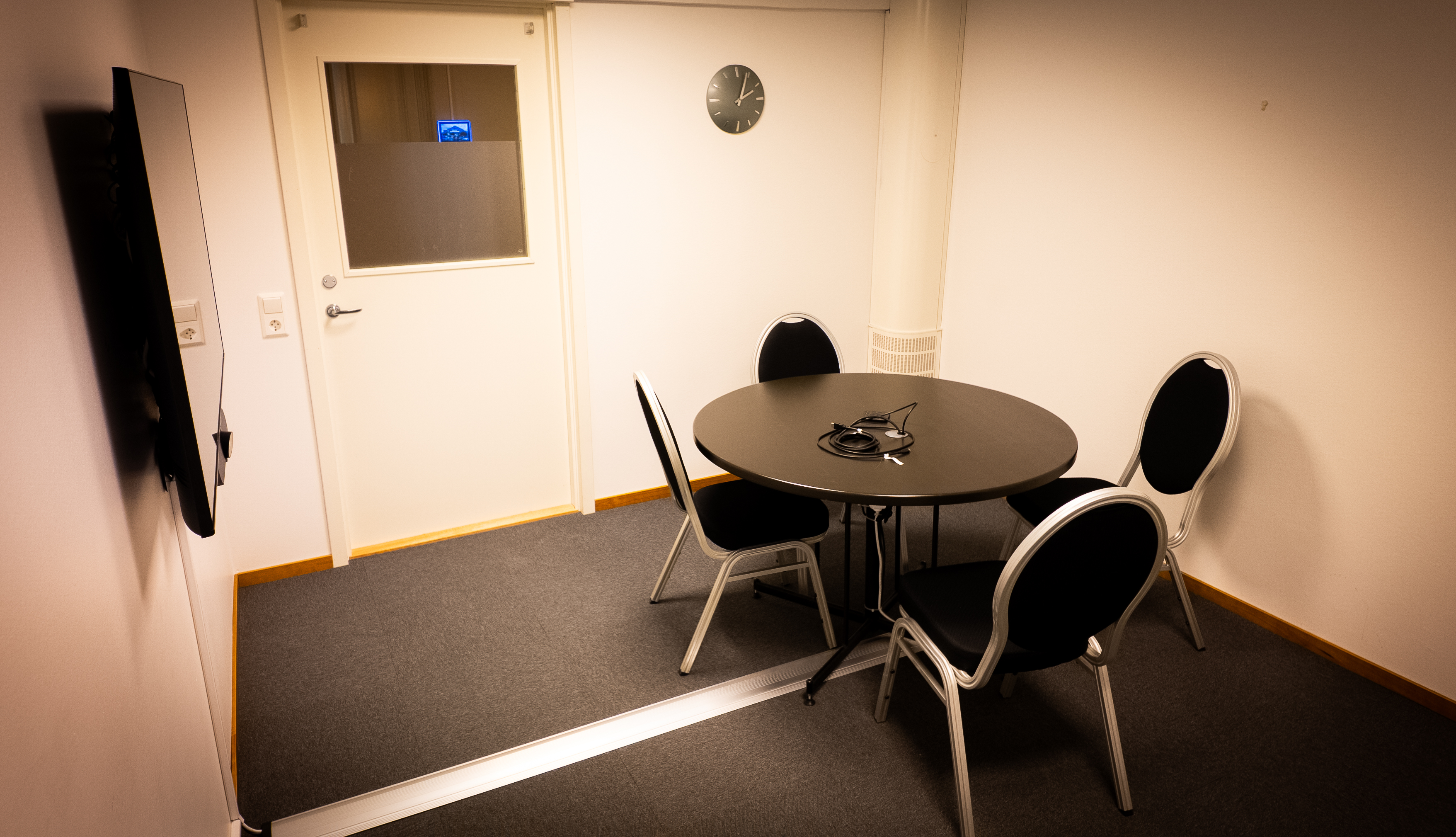 Small conference room with a round table, four chairs, wall clock, and television screen.