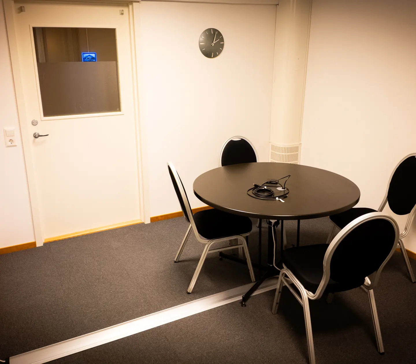 Litet mötesrum med runt bord och fyra stolar, väggklocka och stängd dörr.