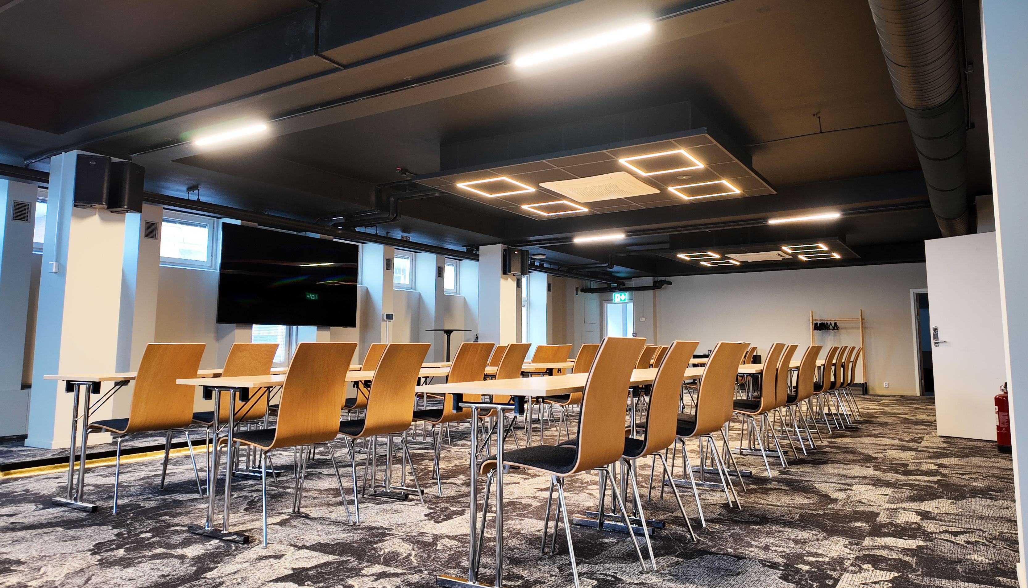 Modern conference room with rows of wooden chairs and tables, featuring a large screen and contemporary ceiling lights.