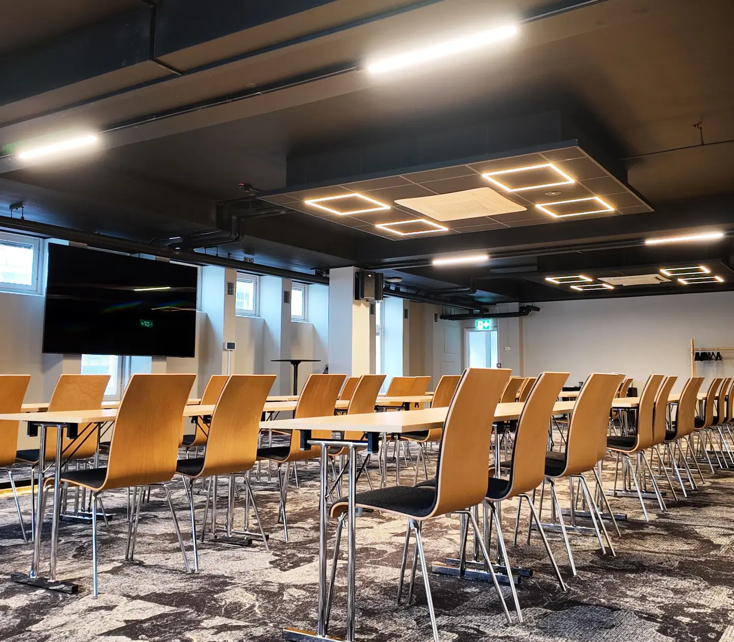 Modern konferensrum med ljusa trästolar och bord, takmonterad belysning och stor tv-skärm på väggen.