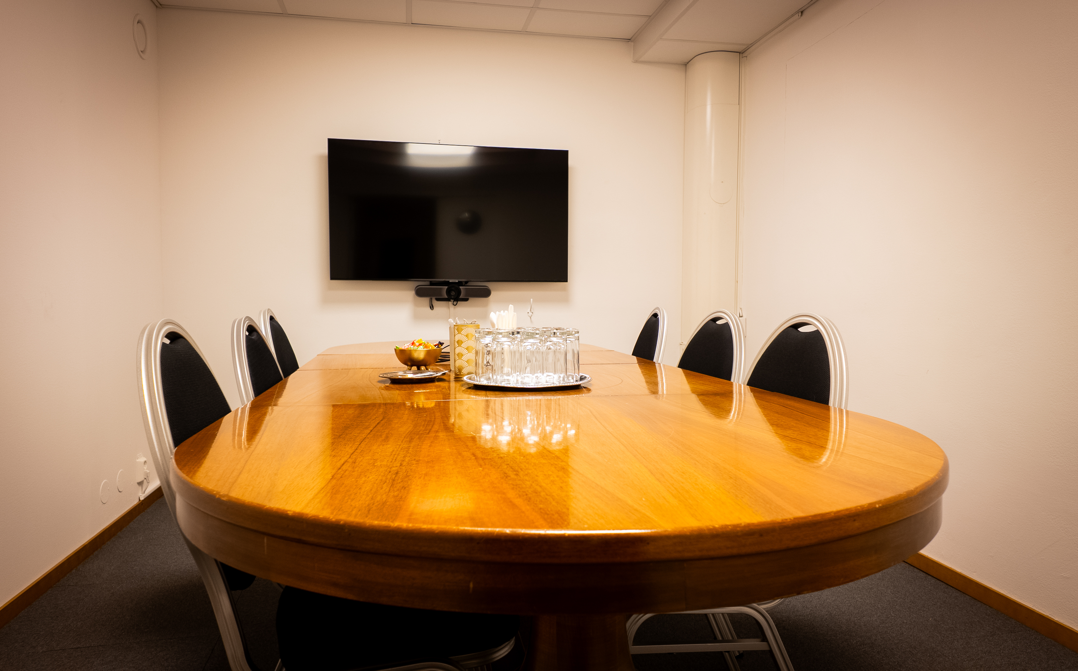 Modern conference room with a wooden oval table, six black chairs, a wall-mounted flat-screen TV, and a set of glasses and snacks in the center.