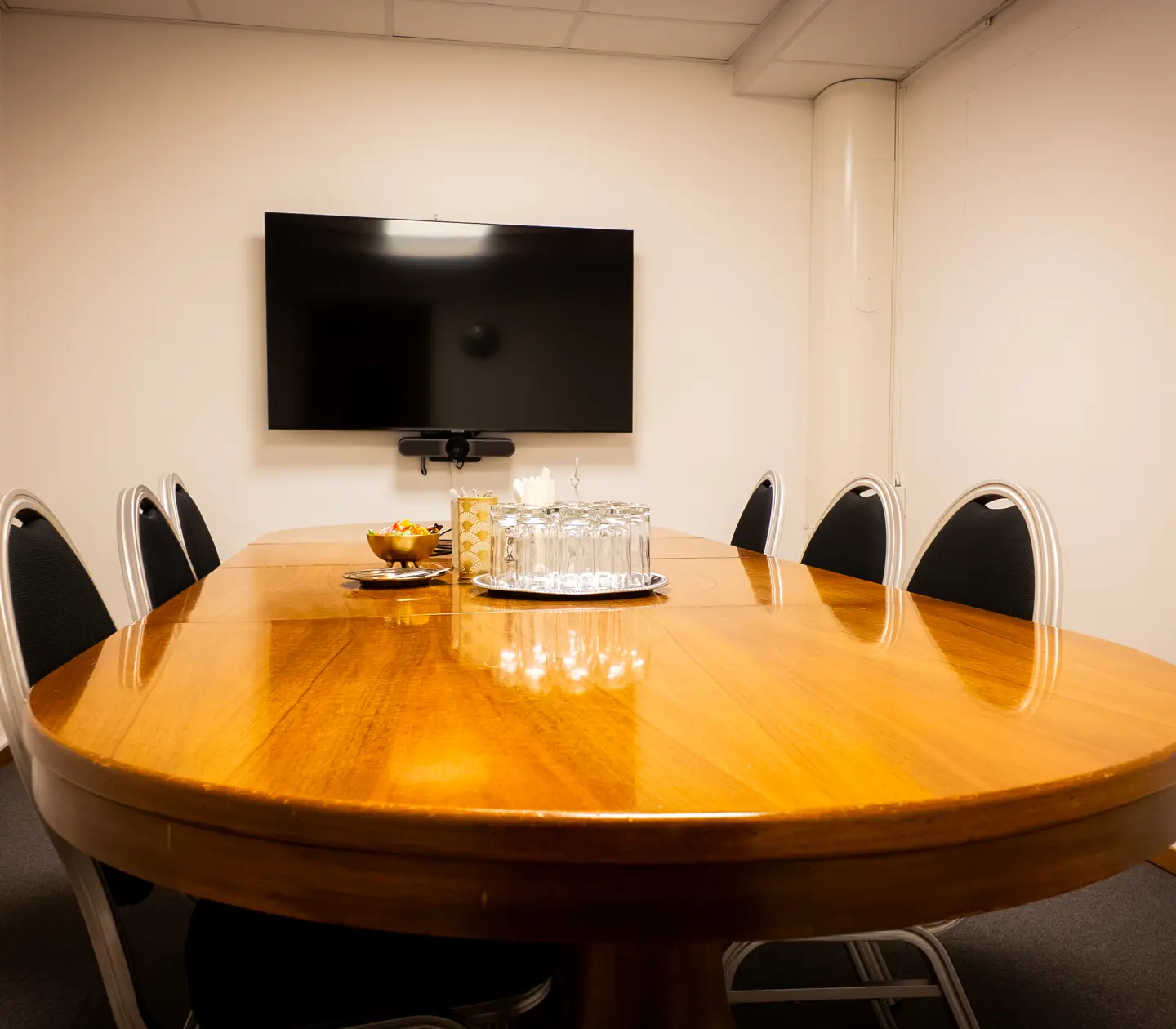 Ovalt konferensbord med svarta stolar, en TV på väggen, glas och karaffer, i ett mötesrum.