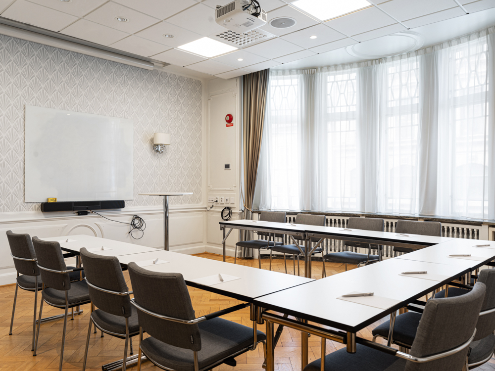 Konferensrum med flera bord och stolar uppställda i rader, ljusa väggar och parkettgolv, stora fönster med långa gardiner.