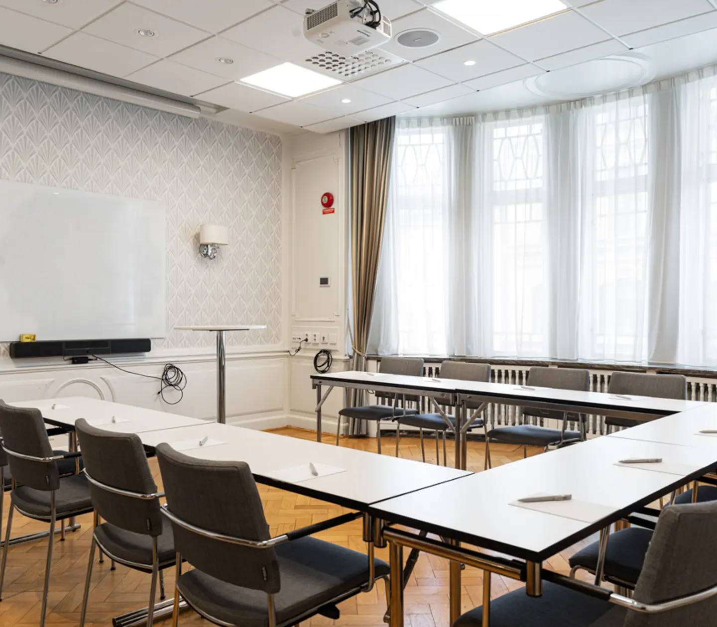 Konferensrum med flera bord och stolar uppställda i rader, ljusa väggar och parkettgolv, stora fönster med långa gardiner.
