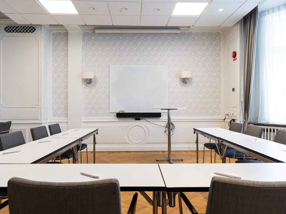 Konferensrum med U-formade bord, stolar, projektorskärm och fönster som släpper in naturligt ljus.