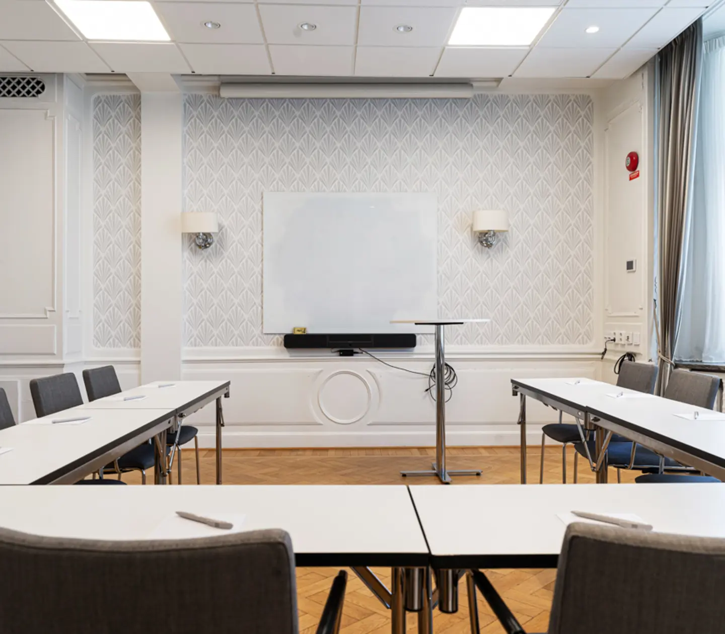 Konferensrum med U-formade bord, stolar, projektorskärm och fönster som släpper in naturligt ljus.