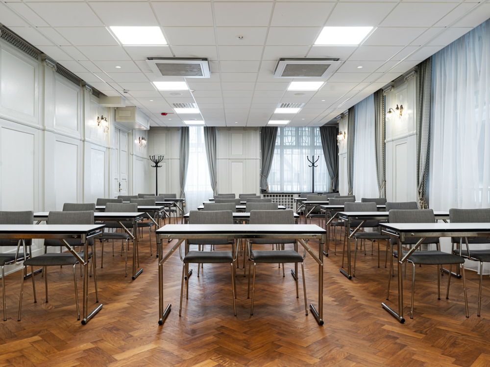 Tomt konferensrum med gråa stolar och bord på ett mörkt trägolv, vita väggar och stora fönster med ljusinsläpp.