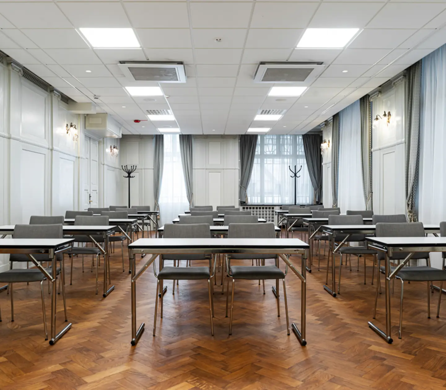 Tomt konferensrum med gråa stolar och bord på ett mörkt trägolv, vita väggar och stora fönster med ljusinsläpp.
