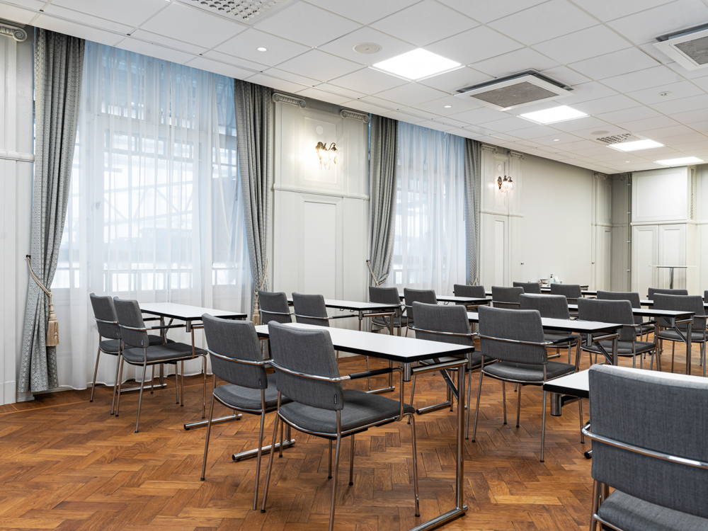 Konferensrum med moderna skrivbord och stolar, träparkettgolv och stora fönster med vida gardiner.