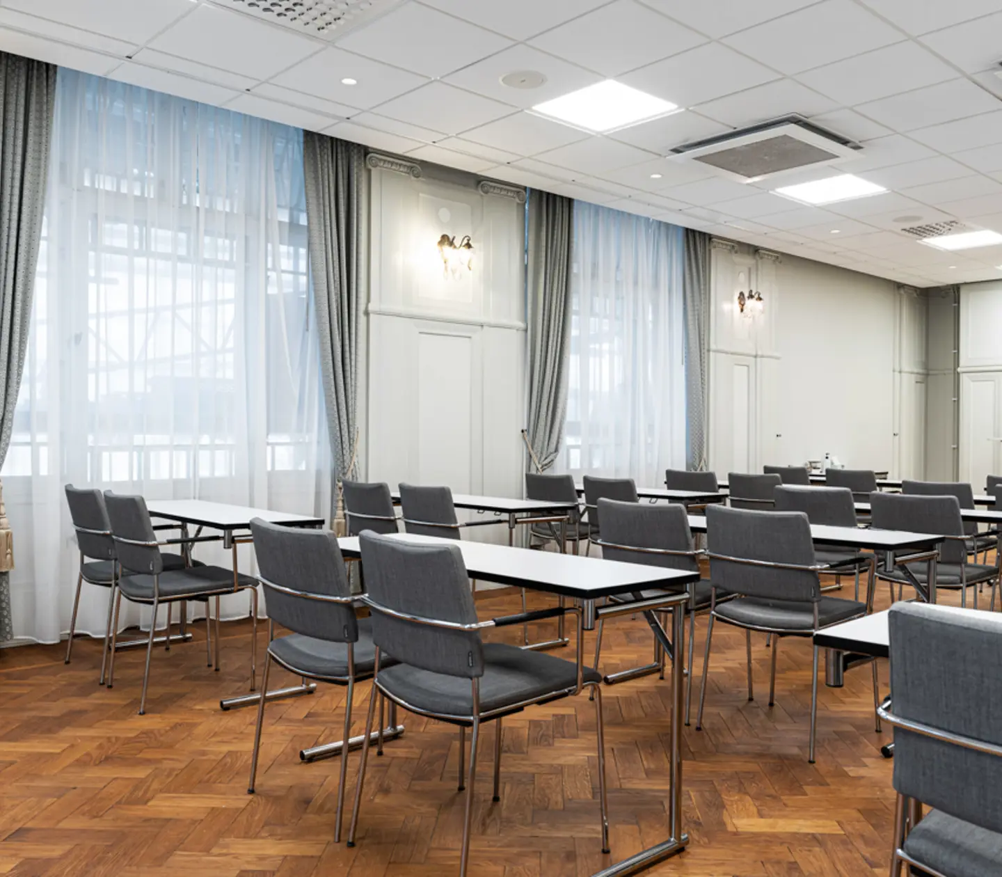 Konferensrum med moderna skrivbord och stolar, träparkettgolv och stora fönster med vida gardiner.