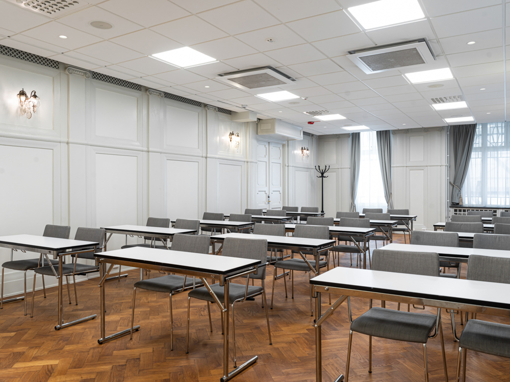 Konferensrum med flera bord och stolar uppställda i rader, ljusa väggar och parkettgolv, stora fönster med långa gardiner.