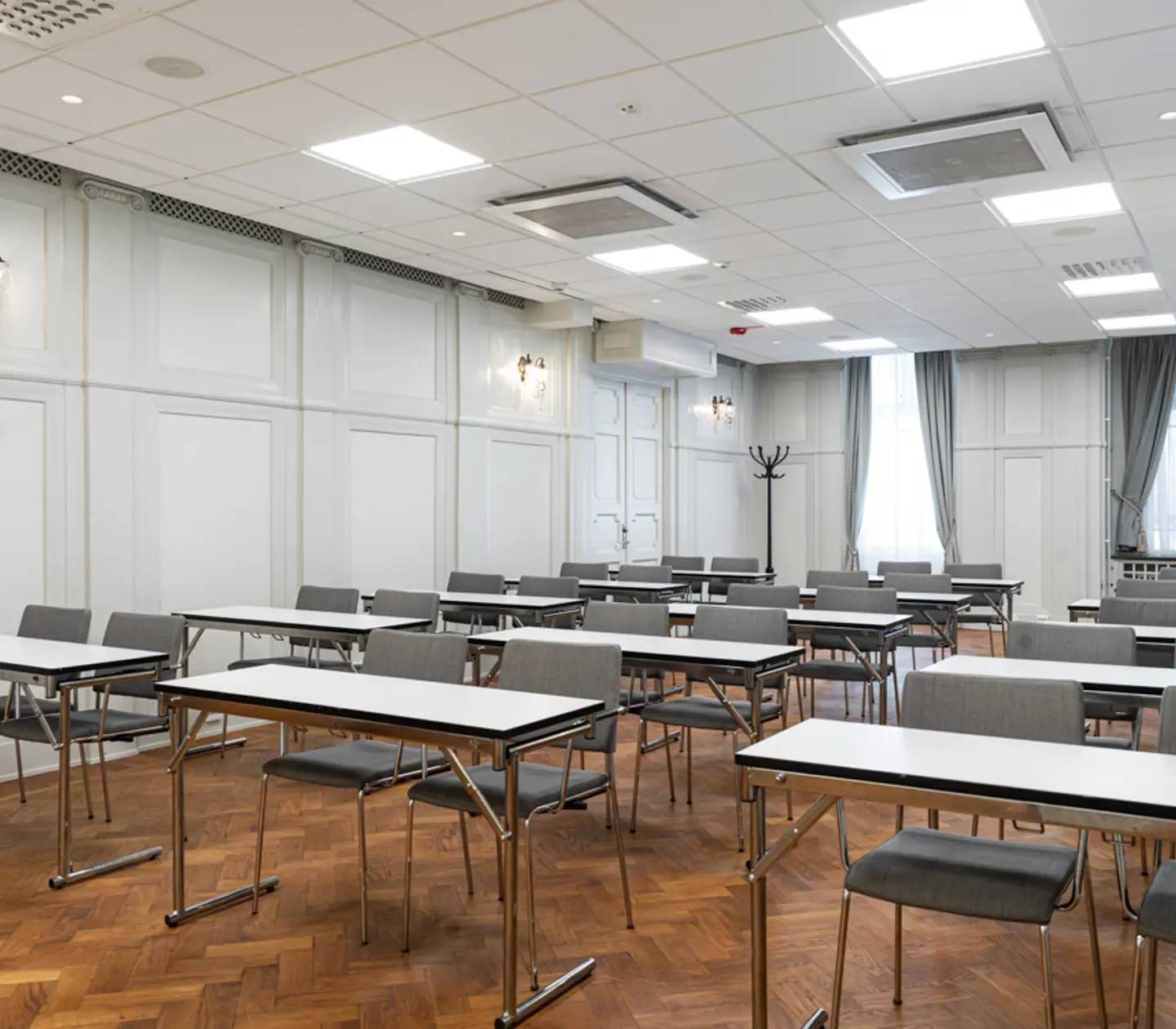 Konferensrum med flera bord och stolar uppställda i rader, ljusa väggar och parkettgolv, stora fönster med långa gardiner.