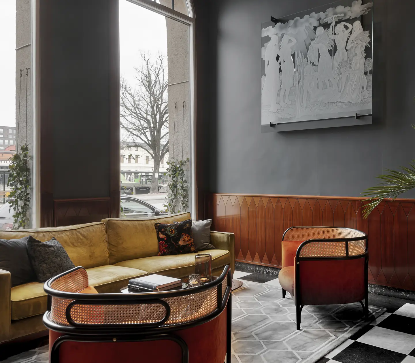 Cozy hotel lobby with modern decor, featuring a yellow sofa, rattan chairs, large windows, and a potted plant against dark gray walls.