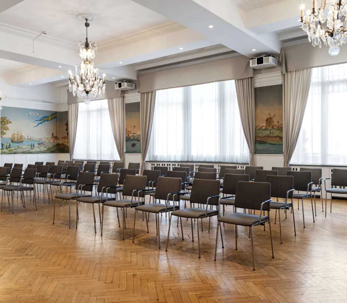 Ljus möteslokal med rad av tomma stolar, elegant parkettgolv och kristallkronor. Väggmålningar och stora fönster med gardiner.