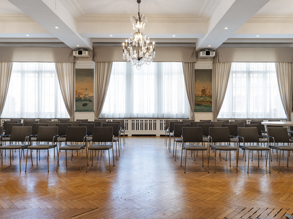 Ljus konferenssal med stolsuppställning, parkettgolv, och elegant kristallkrona.