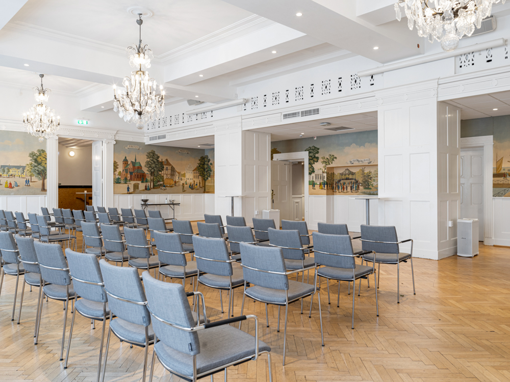 Konferensrum med rader av grå stolar, vackra kristallkronor och väggmålningar, trägolv och ljus inredning.