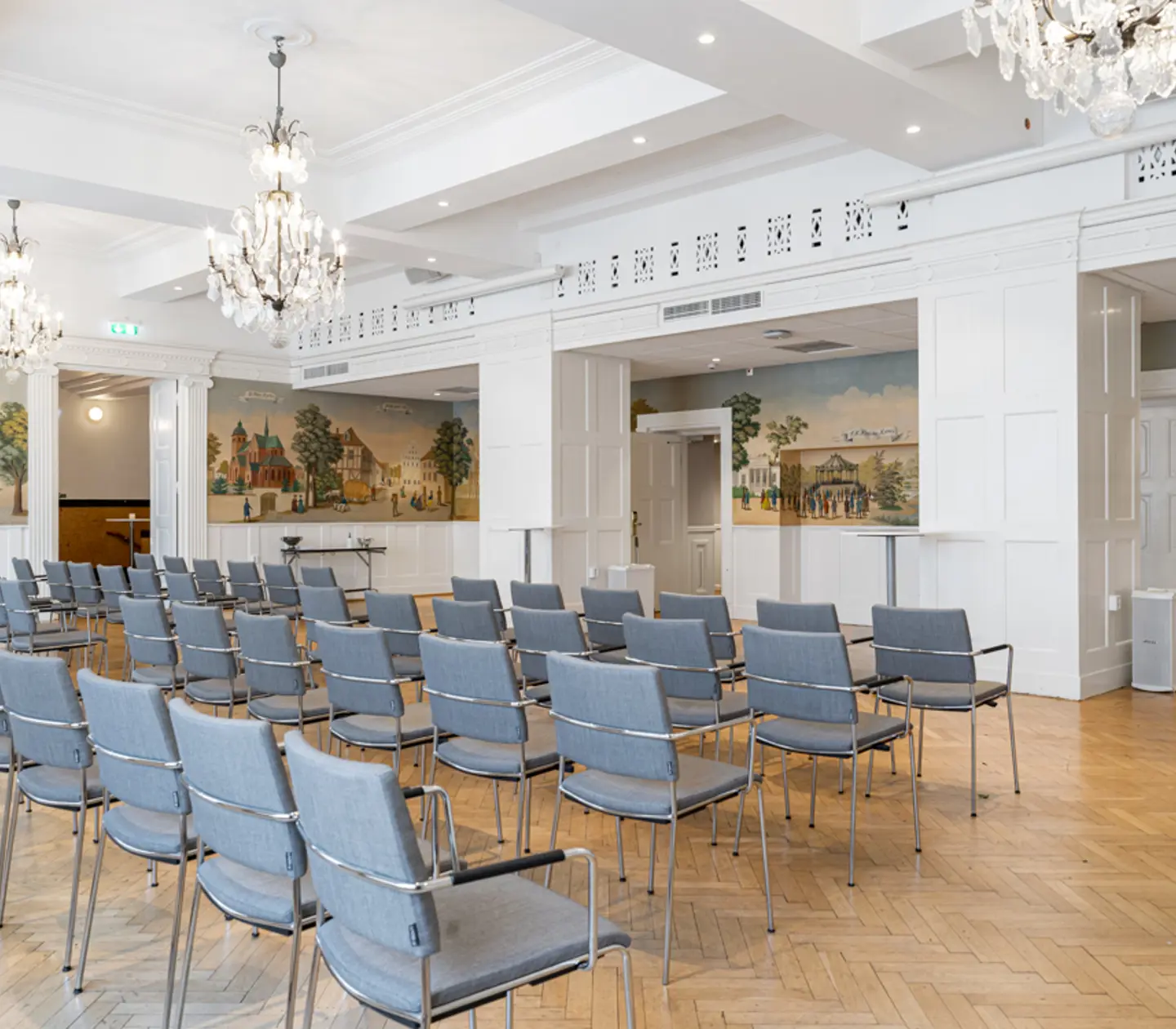 Konferensrum med rader av grå stolar, vackra kristallkronor och väggmålningar, trägolv och ljus inredning.