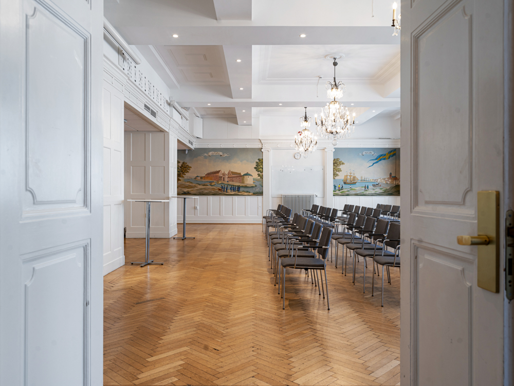 Öppen dörr till konferensrum med rader av stolar, parkettgolv och ljuskronor i taket.