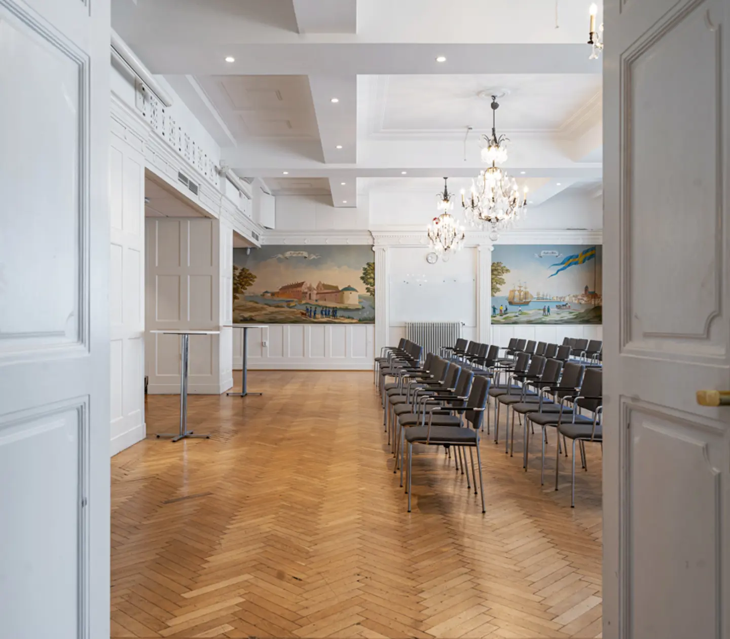 Öppen dörr till konferensrum med rader av stolar, parkettgolv och ljuskronor i taket.
