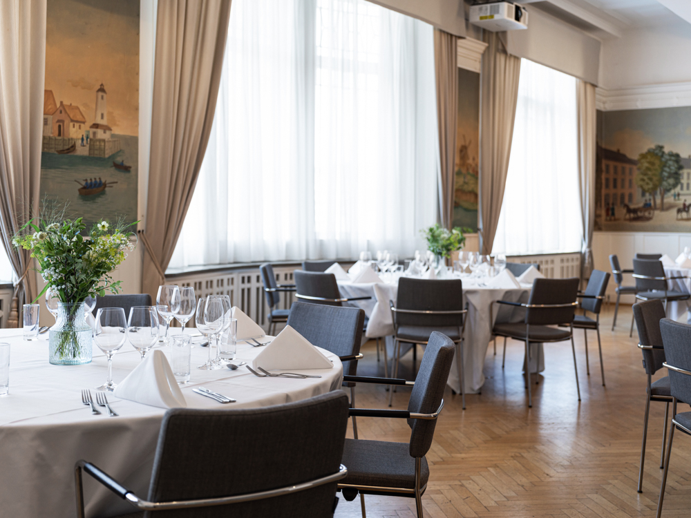 Elegant middagsmiljö med dukade runda bord, blommor i vaser, och smakfull inredning i en ljus sal med stora fönster och målade väggar.